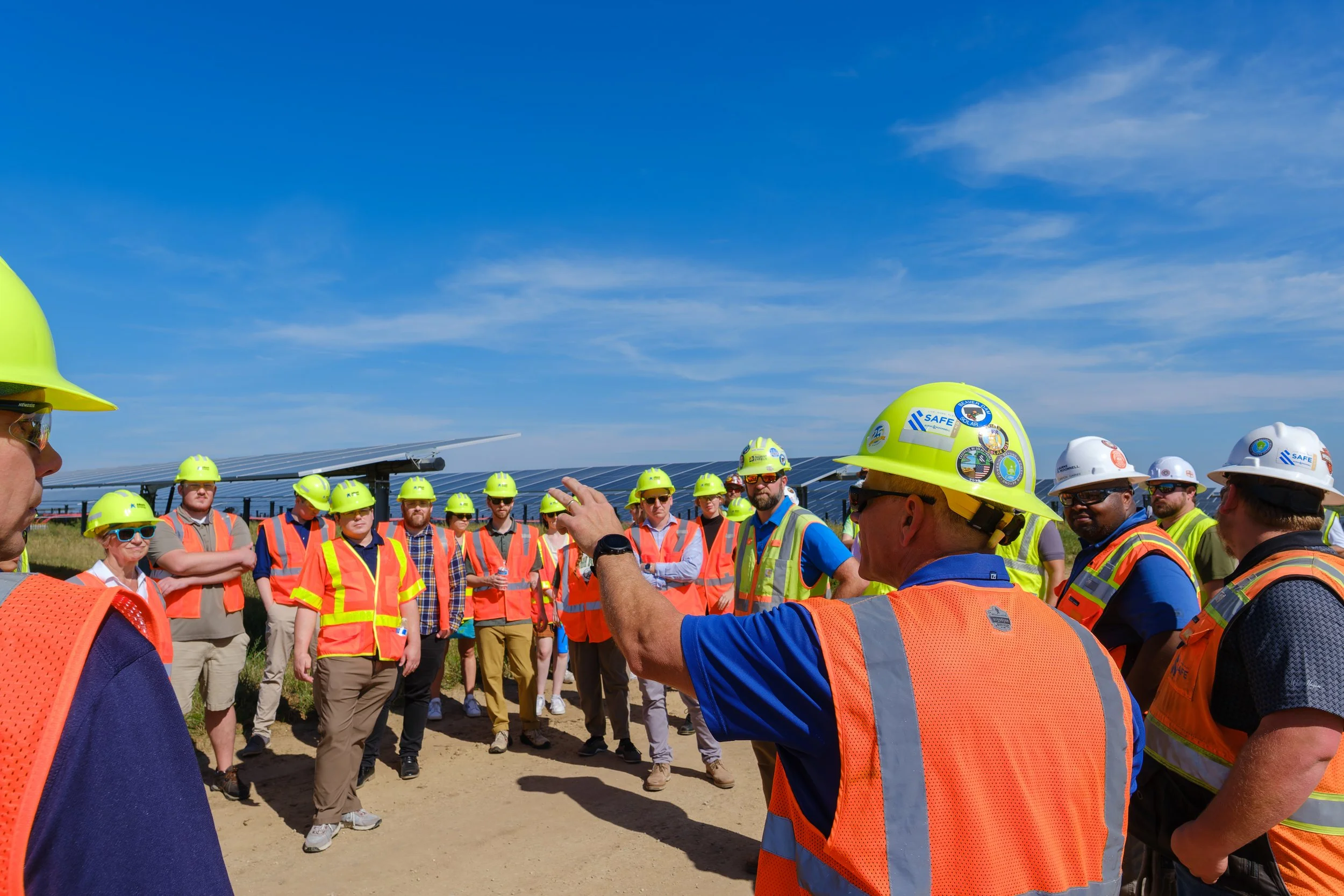 Group of people wearing safety helmets and vests gathered outdoors for a briefing near solar panels on a sunny day.
