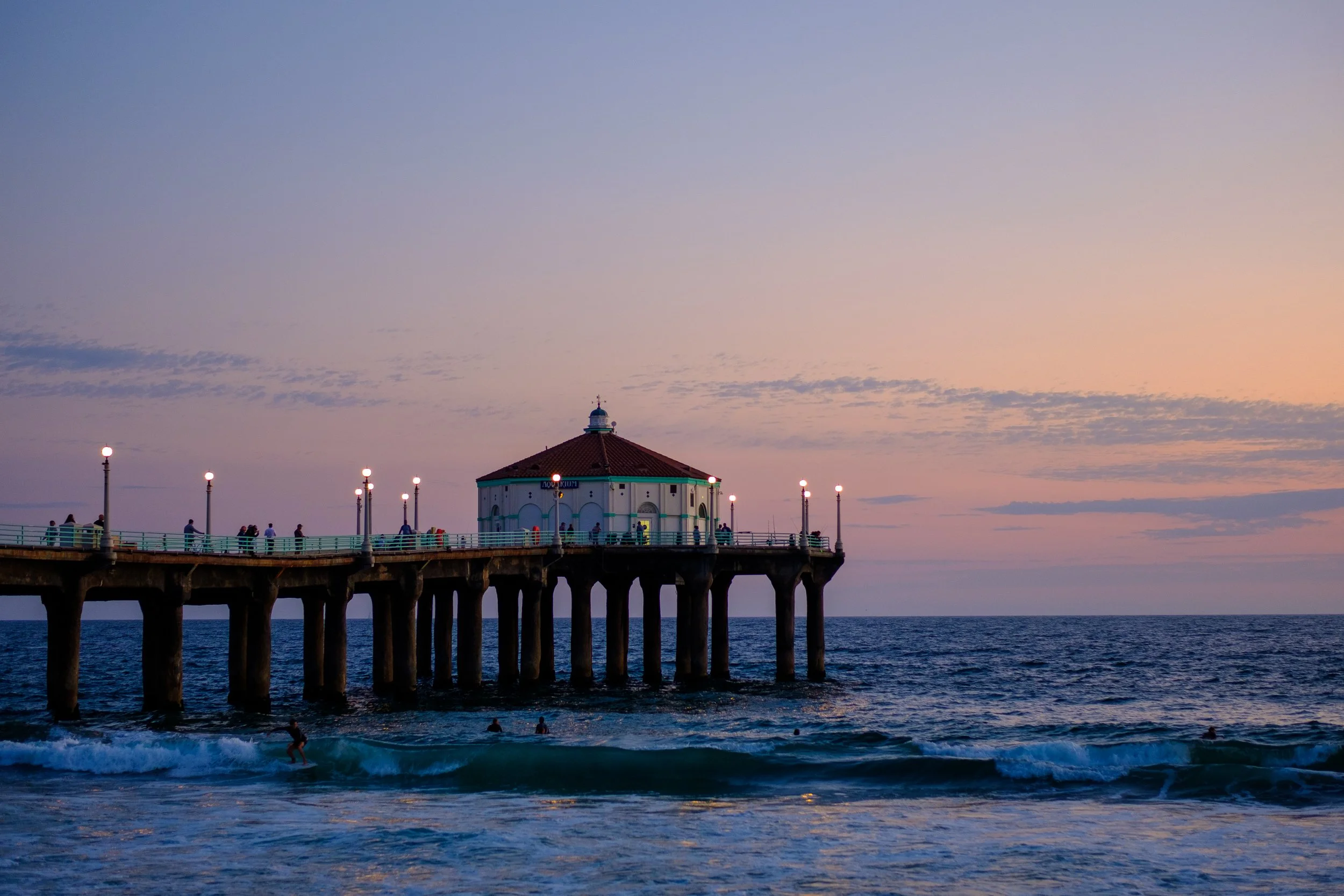 The Manhattan Beach Pier extending into the Pacific Ocean during sunset, with a pavilion at the end and people walking and surfing on the water.