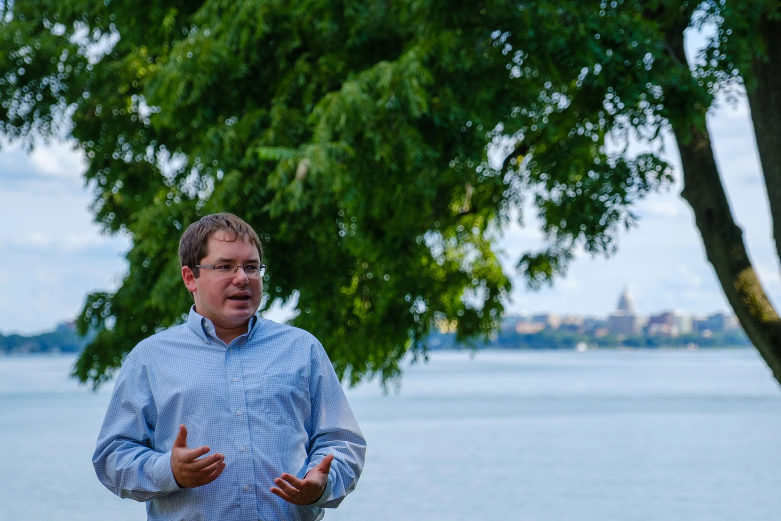 Wisconsin State Senator Mark Spreitzer speaks on the north shore of Lake Mendota, with trees behind him and the Madison skyline in the distance, featuring the Wisconsin State Capitol dome.