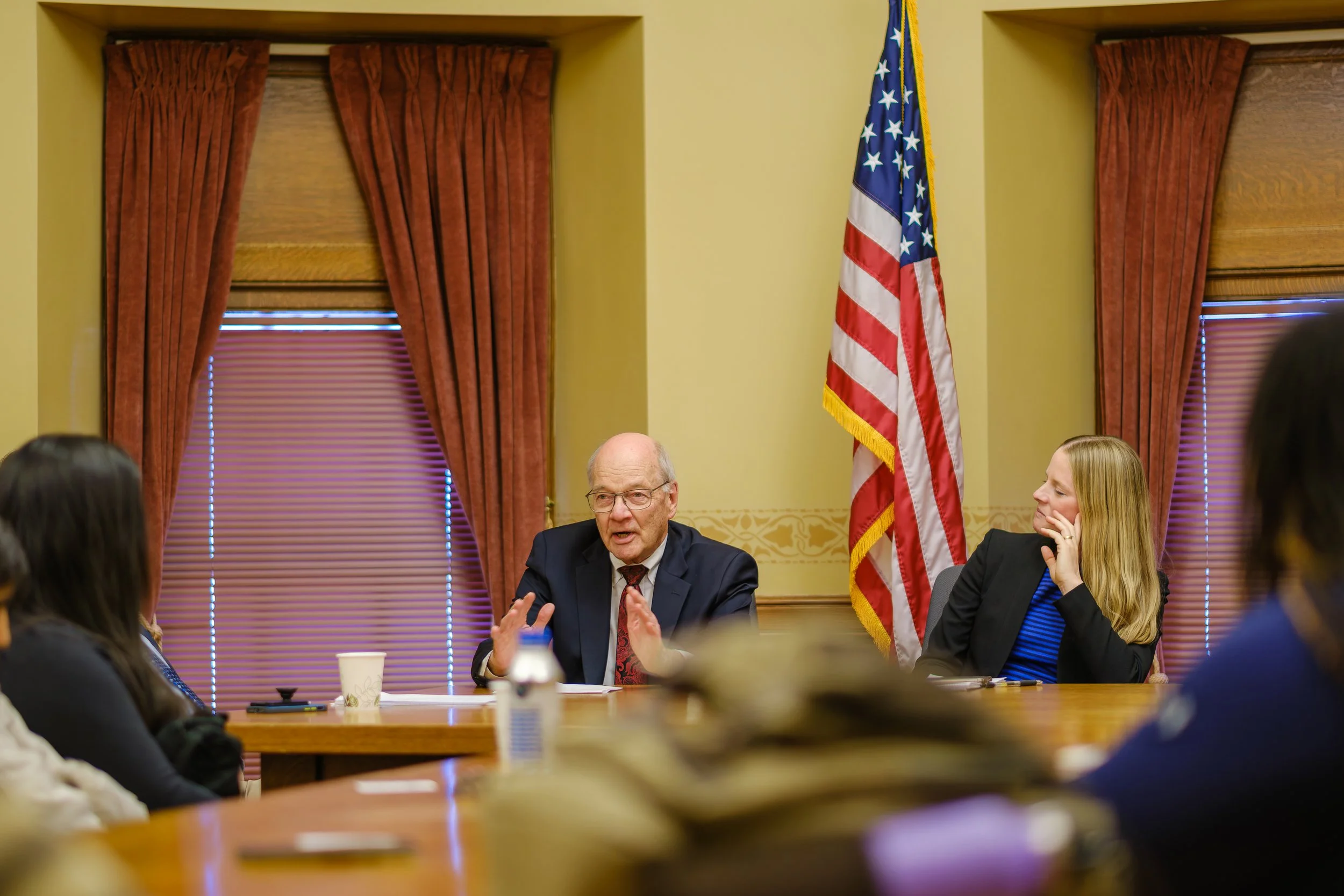 Legislative Fiscal Bureau Director Bob Lang speaks at a meeting in the Wisconsin State Capitol, seated at a wooden table with several people. An American flag and red curtains are in the background.