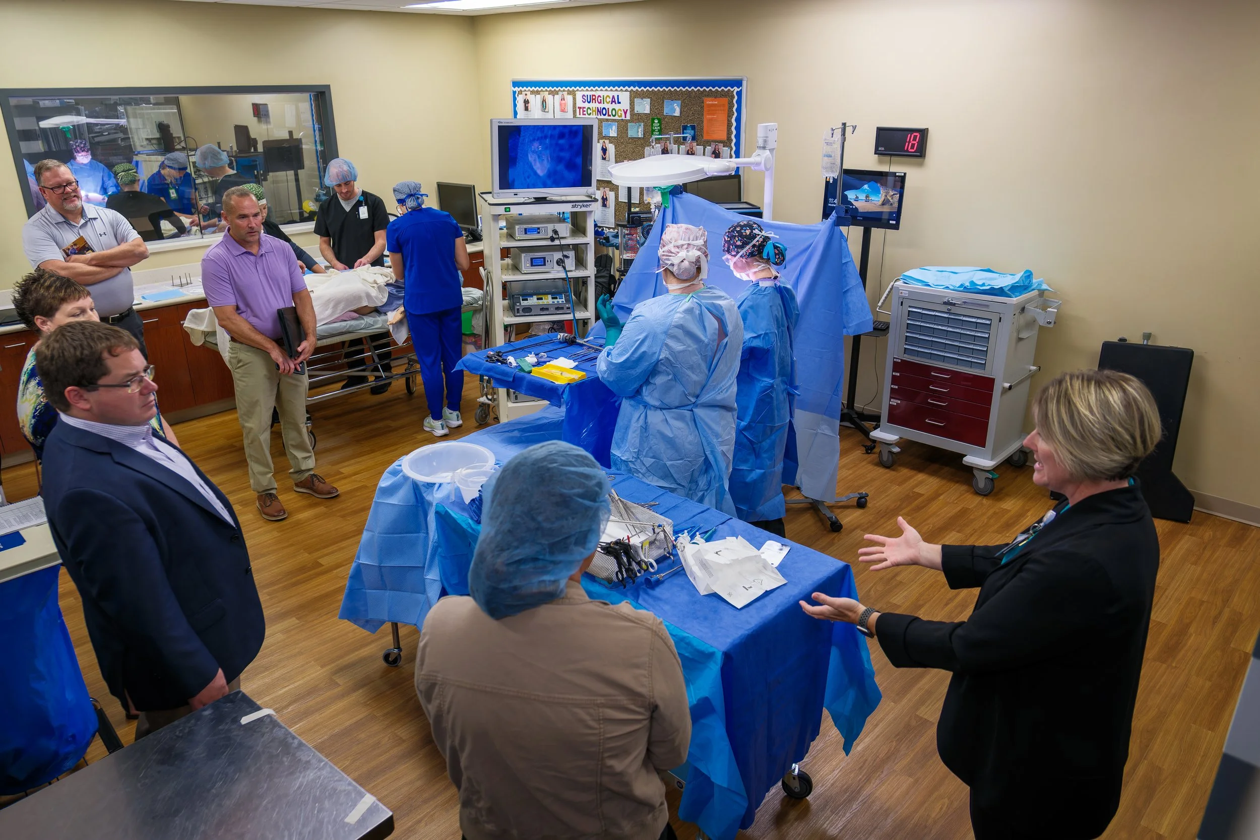 Medical professionals and students in a hospital operating room during a training, with some preparing surgical instruments and others observing, and a patient on a stretcher in the background.