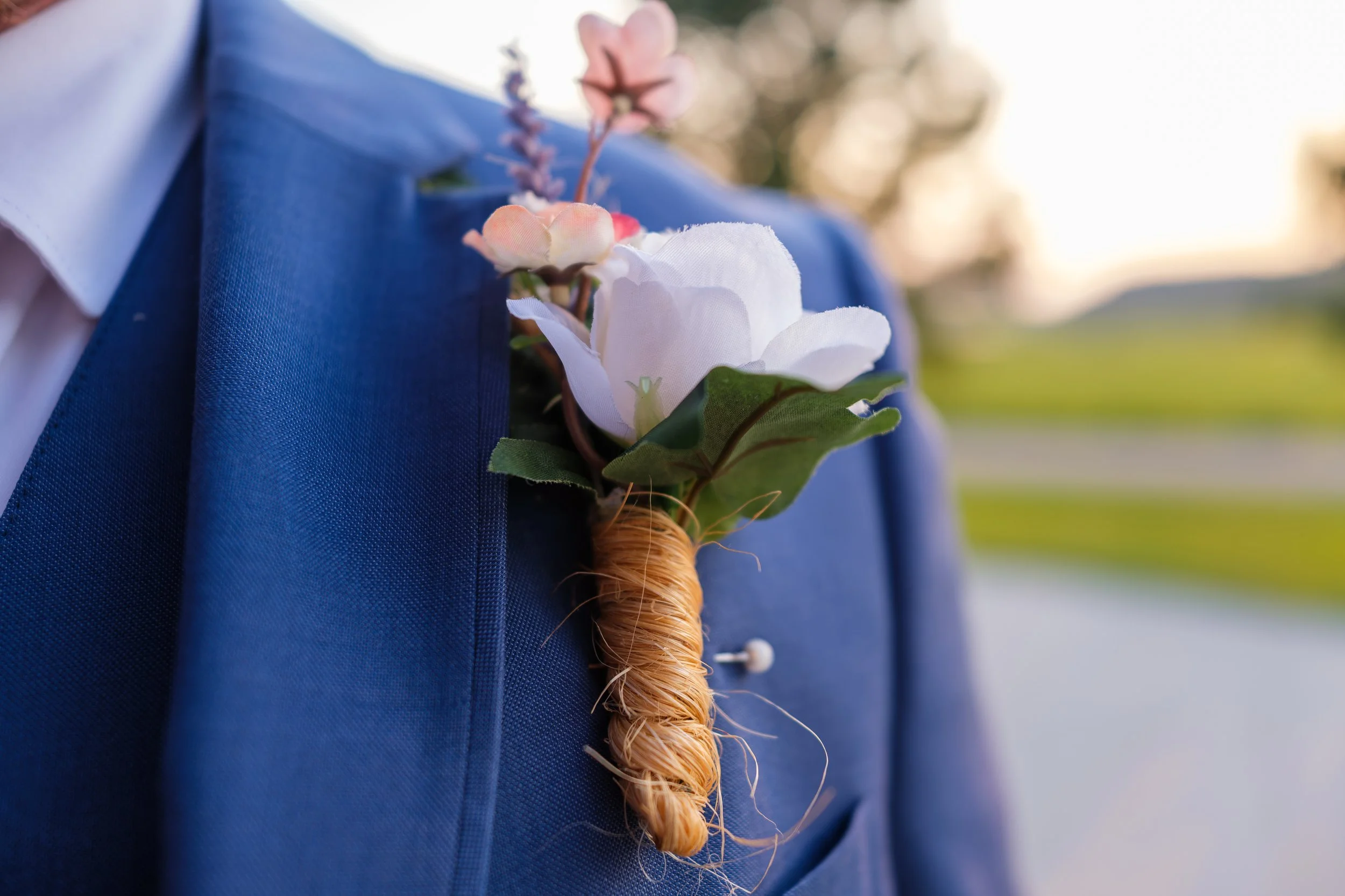 A flower boutonniere with white flowers, green leaves, and a brown wrapped stem is pinned to a blue suit jacket.