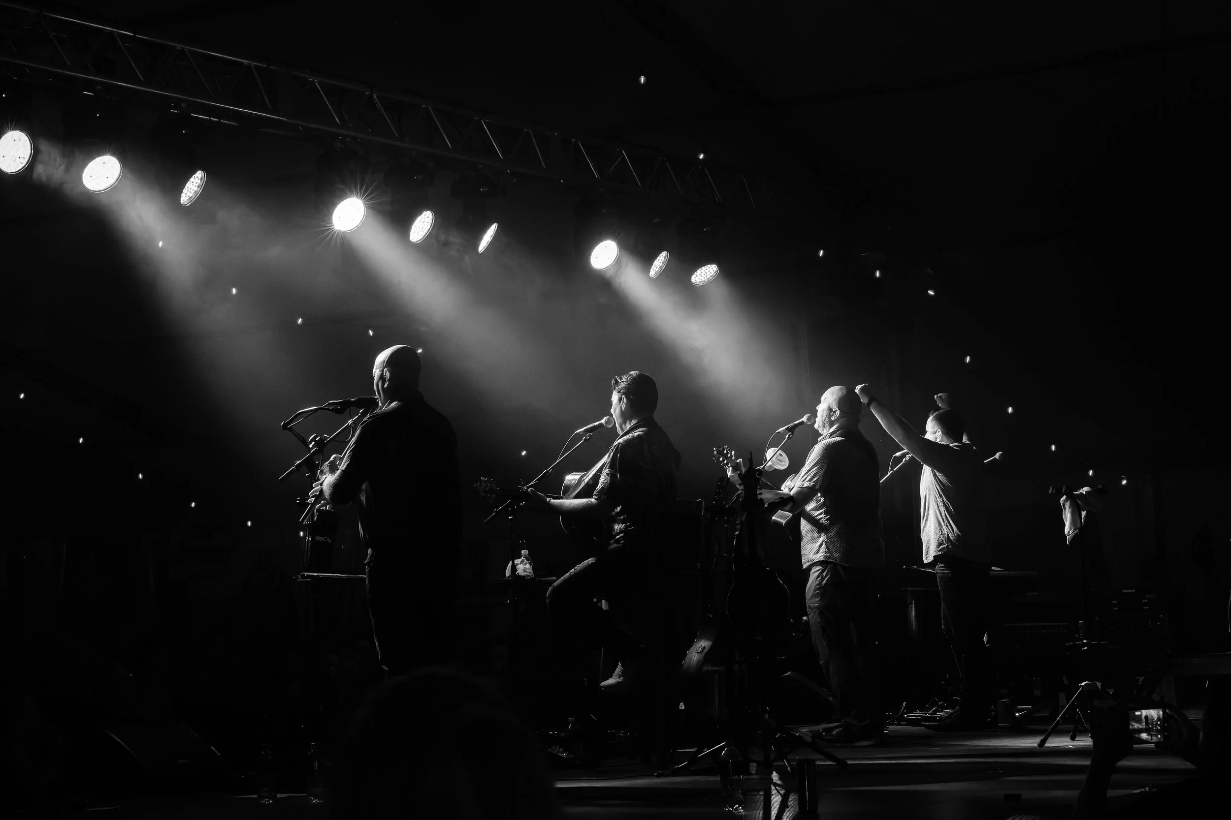 Black and white photo of a music performance by The High Kings with four musicians on stage, illuminated by stage lights, with some instruments visible.