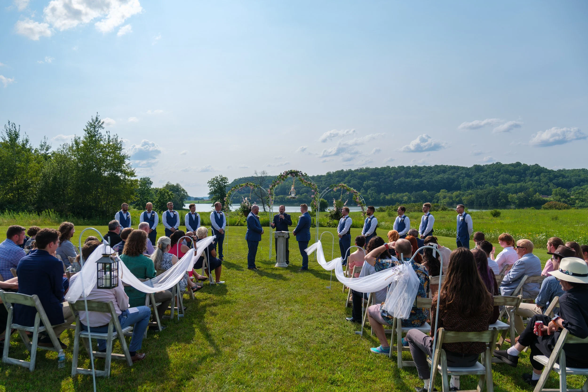 Outdoor wedding ceremony on a grassy field near a lake with green hills in the background. Guests seated on white chairs decorate with white fabric, and the wedding party stands before a floral arch with the officiant in the center. The sky is partly