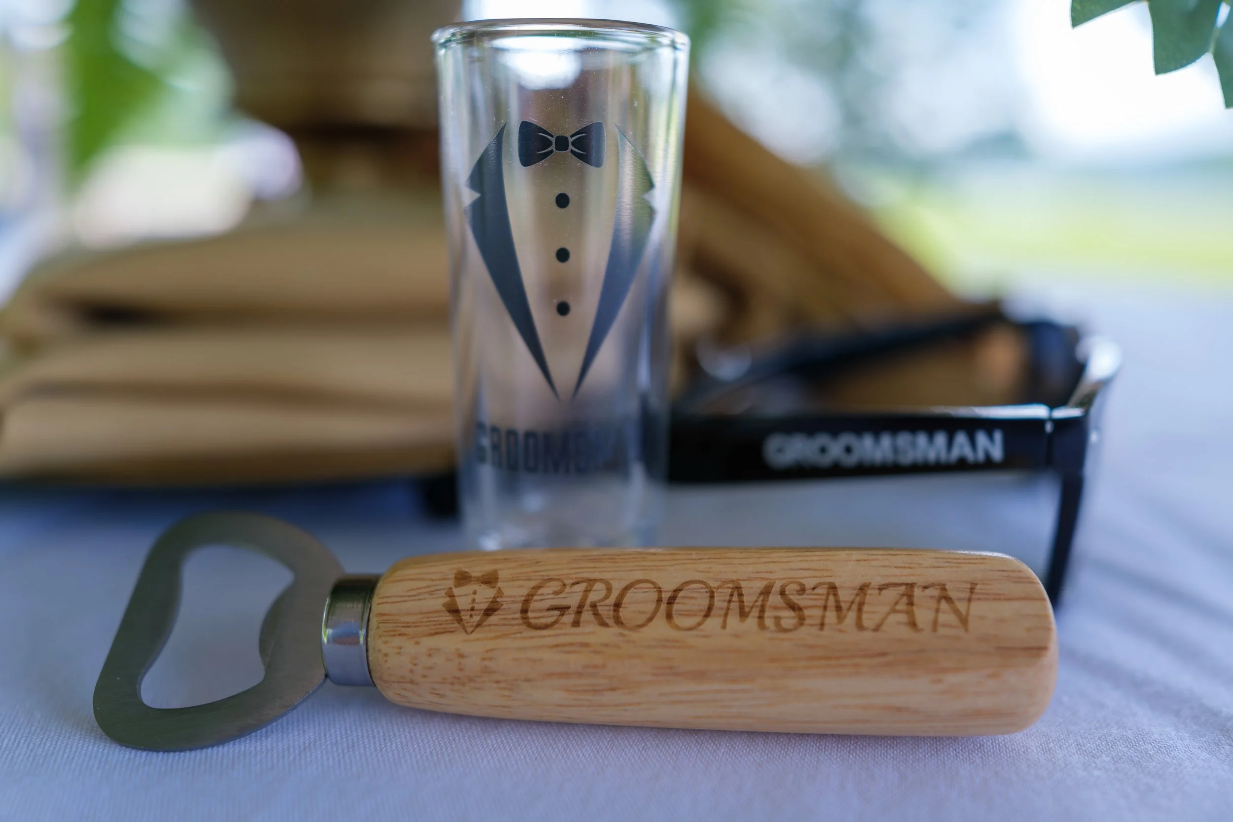 A wooden-handled bottle opener, a tall glass with a tuxedo illustration, and a pair of black sunglasses with 'GROOMSMAN' written on the temple, all on a white surface.