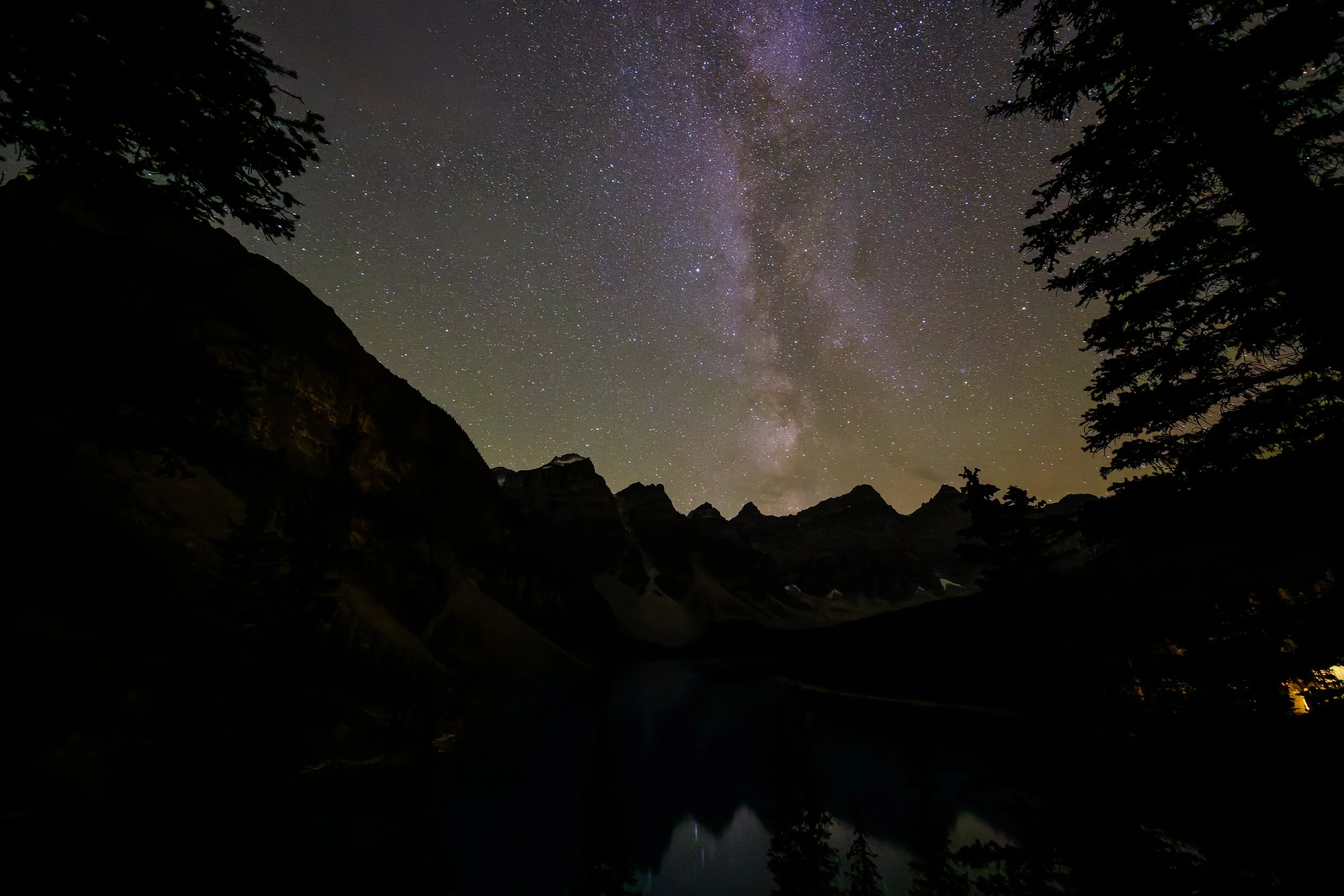 Nighttime landscape of Moraine Lake with silhouetted mountains, trees, and a body of water, under a star-filled sky with the Milky Way galaxy visible in Banff National Park.