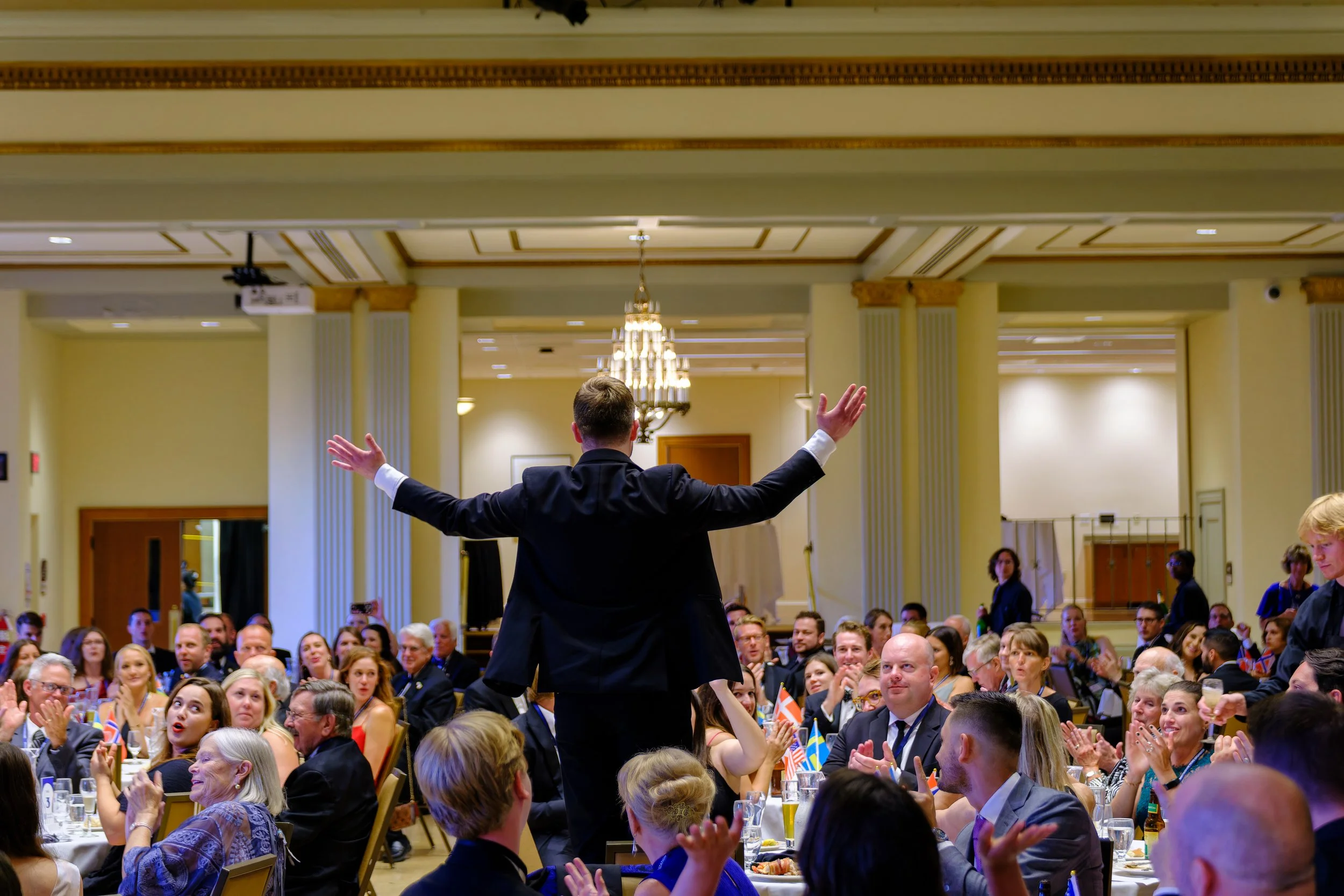 A man in a black suit stands at a banquet, addressing a seated audience with arms outstretched, in a decorated ballroom with chandeliers and columns.