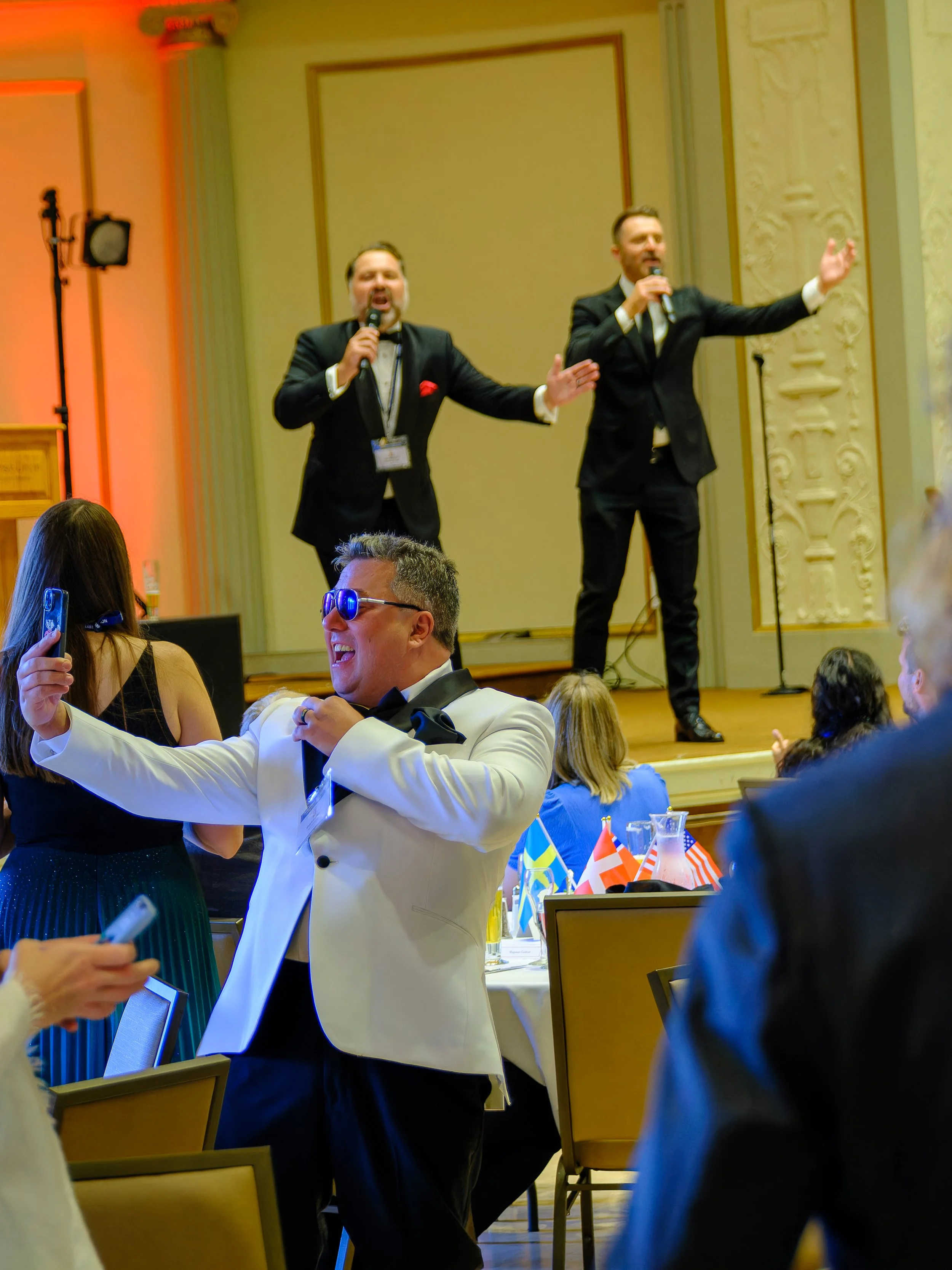 Two men in tuxedos performing on a stage at a formal event with an audience, some of whom are taking photos and enjoying the entertainment.