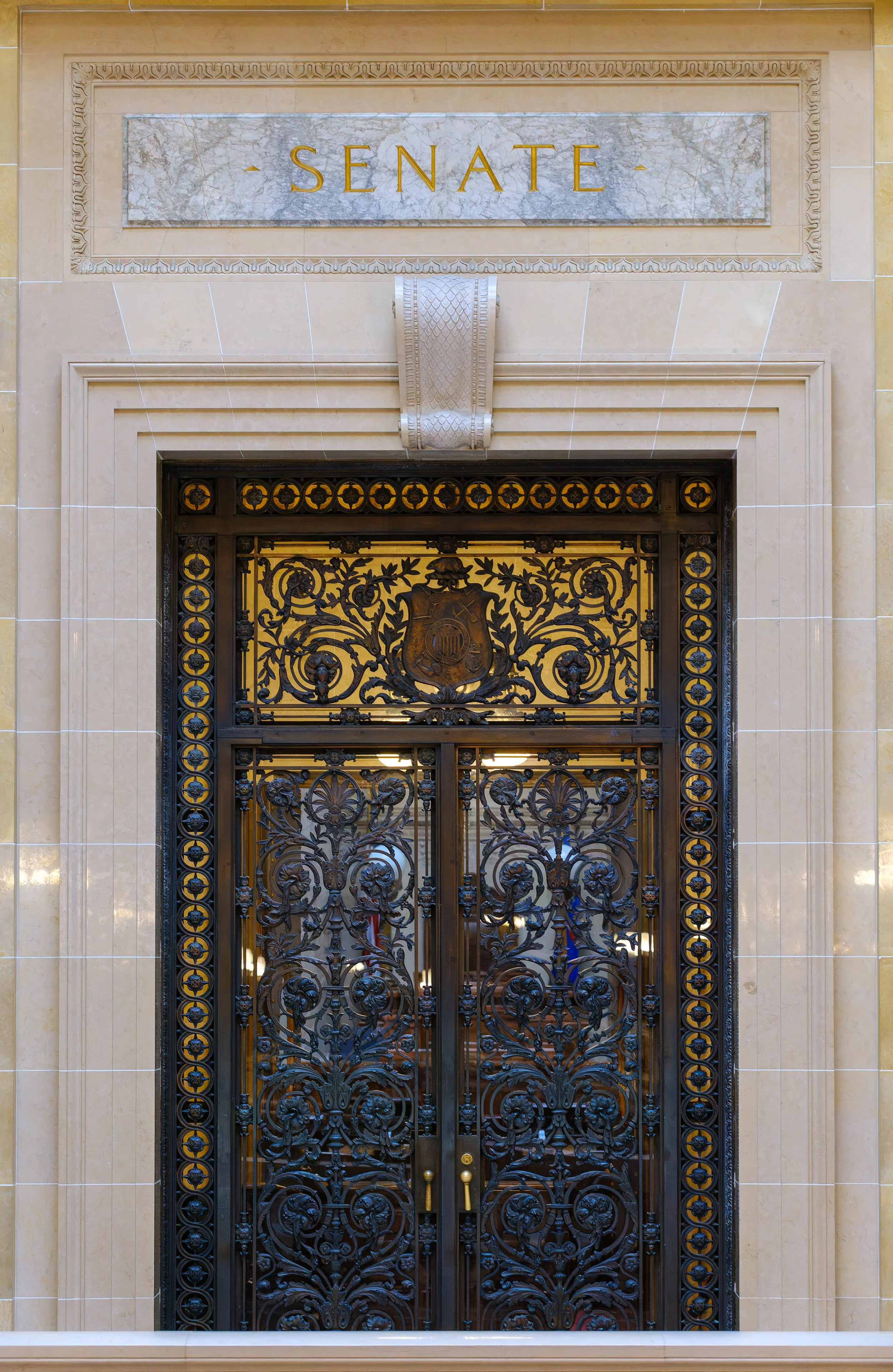 Decorative black iron doors with intricate floral and crest designs, framed by beige marble walls, with an engraving above reading 'Senate' in gold lettering. These doors lead to the floor of the Wisconsin State Senate.