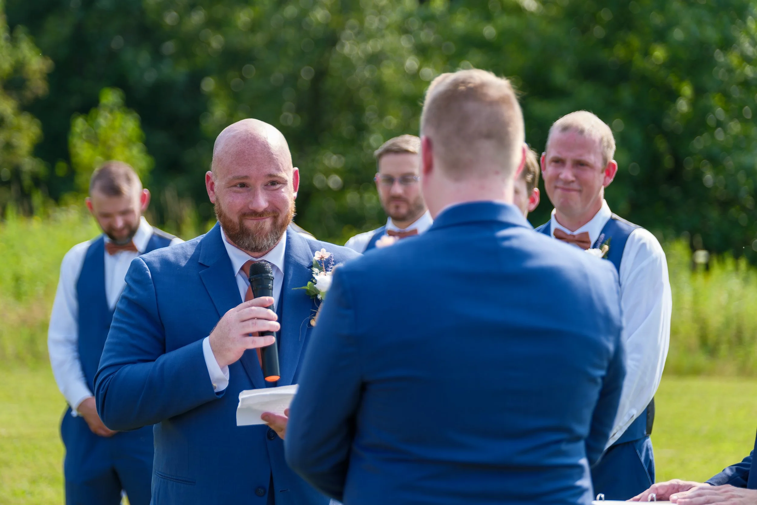 Groom giving a speech during a wedding ceremony outdoors, surrounded by groomsmen, with greenery in the background.