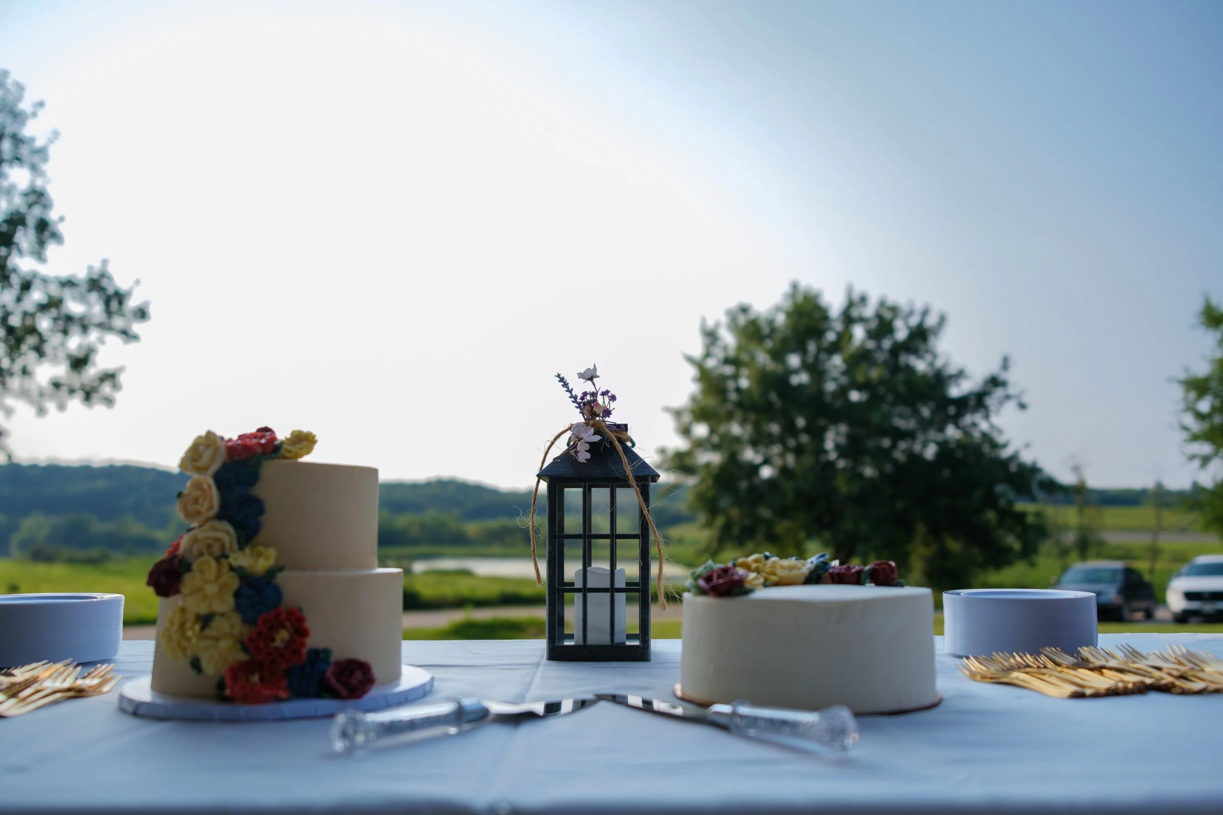Table with two white wedding cakes decorated with colorful flowers, a black lantern in the center, and gold utensils, set outdoors with trees and cars in the background.
