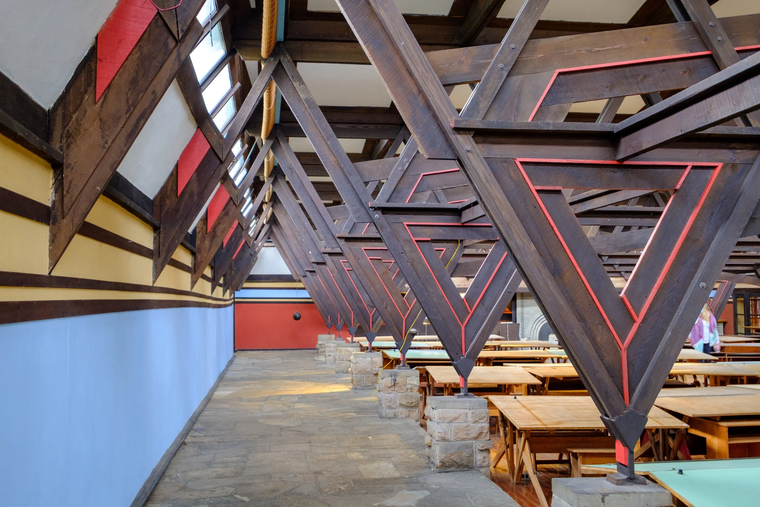Interior of a building at Frank Lloyd Wright's Taliesin in Wisconsin with a sloped wooden ceiling supported by wooden beams with red accents, stone pillars, wooden tables and benches, and blue and red wall sections.