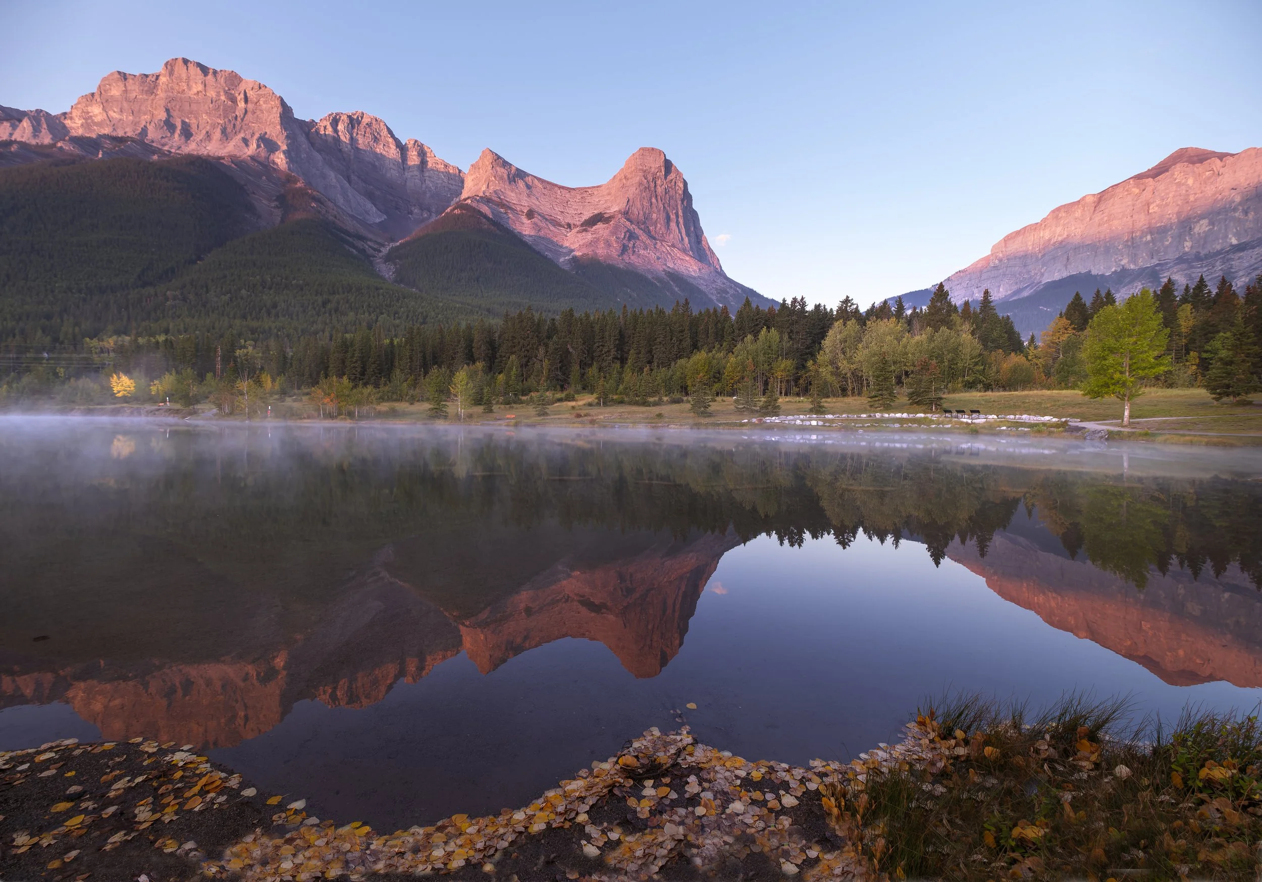 A peaceful mountain landscape with a calm Quarry Lake reflecting the mountains and trees, early morning mist rising from the water, and a clear sky with pink hues from sunrise in Canmore, Alberta.
