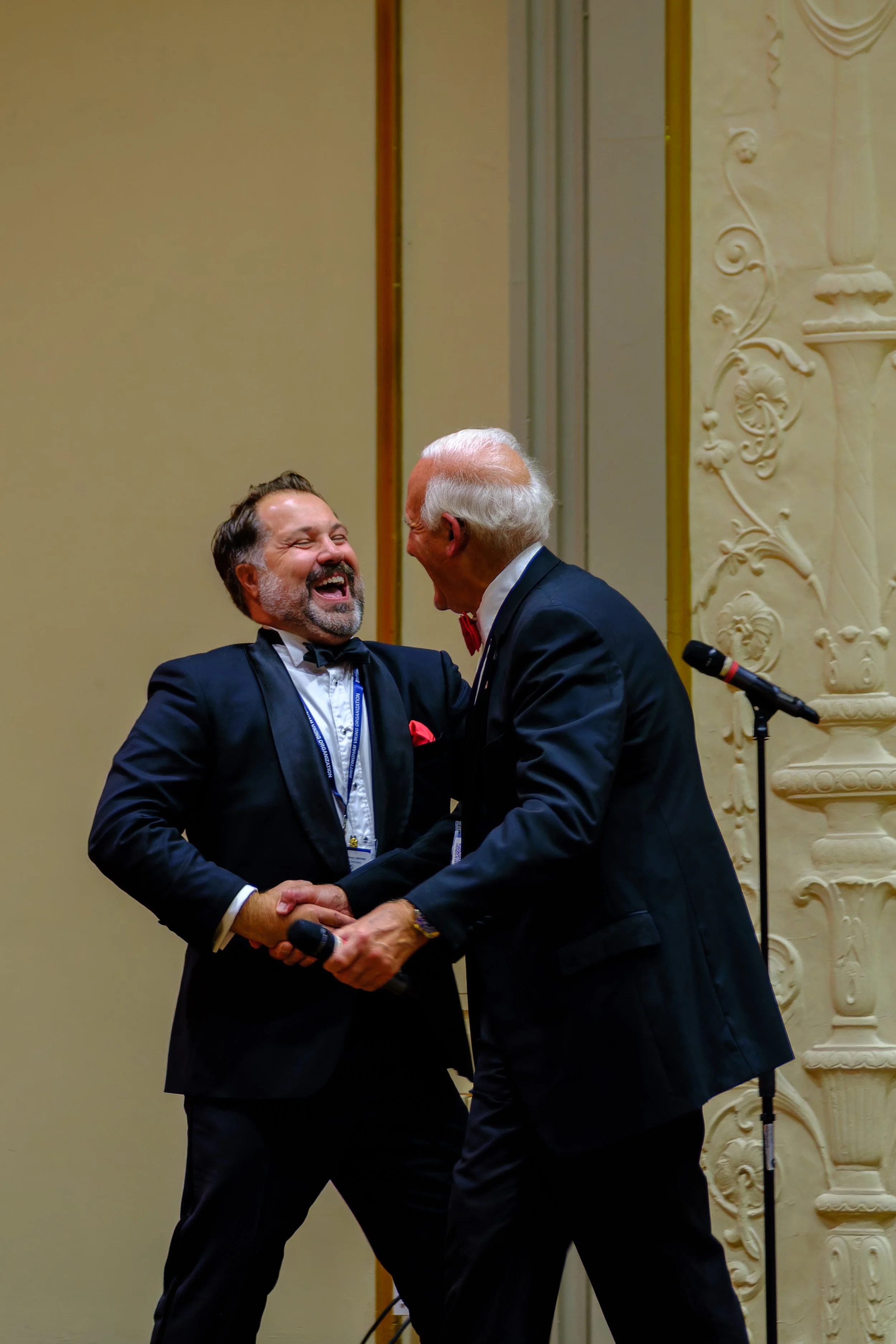 Two men in tuxedos laughing and shaking hands on stage at an event, with microphones nearby.