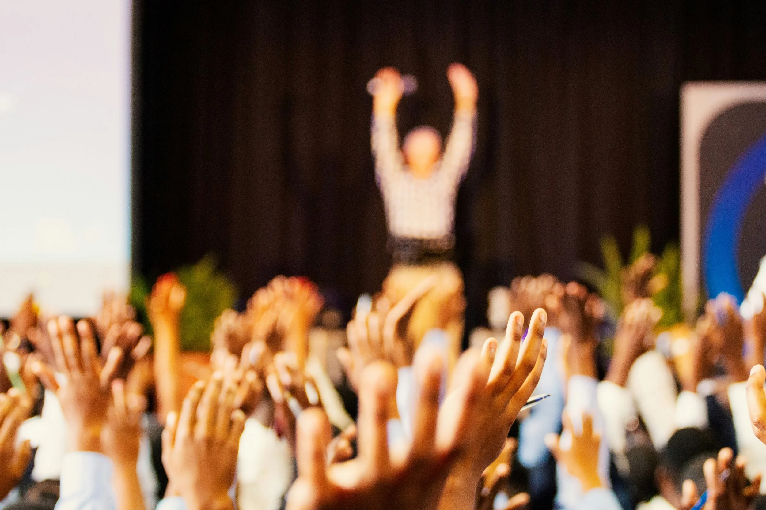 A large crowd of people with raised hands reaching towards a stage where a blurred figure with outstretched arms is standing in front of a dark curtain.
