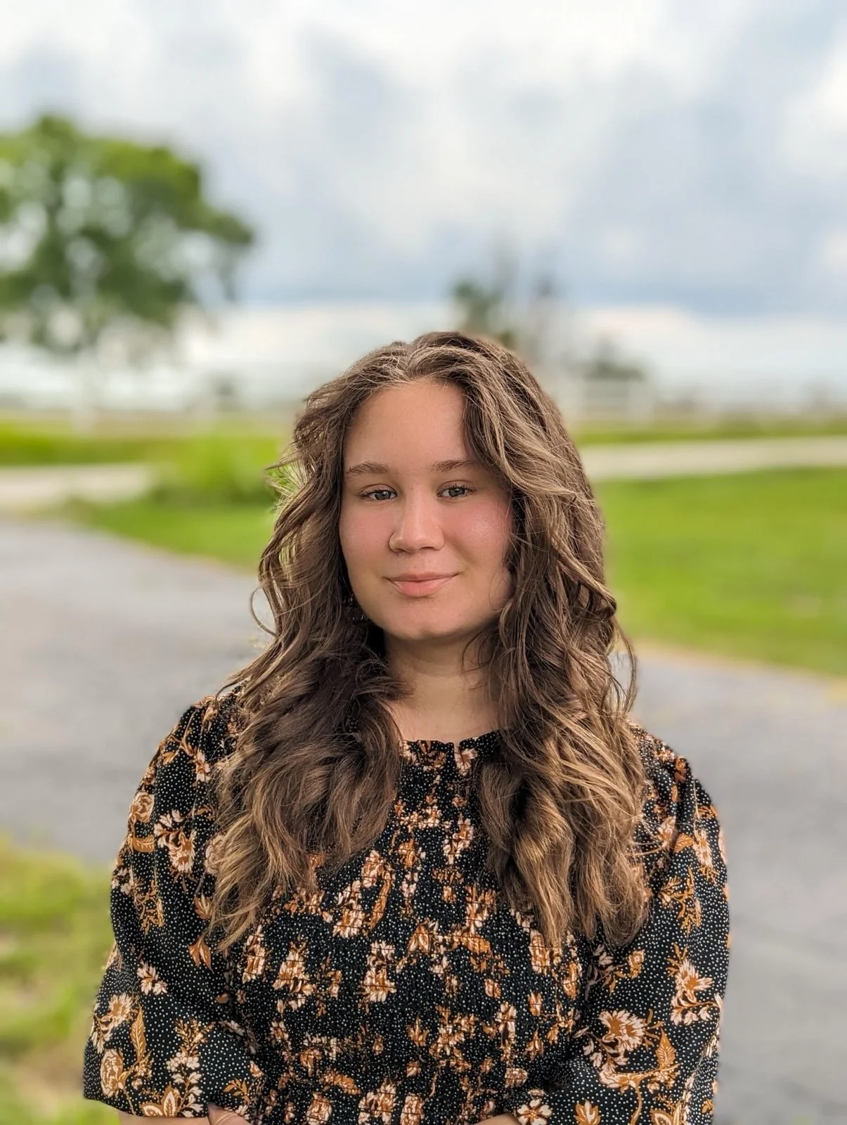 A young woman with long, wavy brown hair posing outdoors on a cloudy day, with greenery and a pathway in the background.