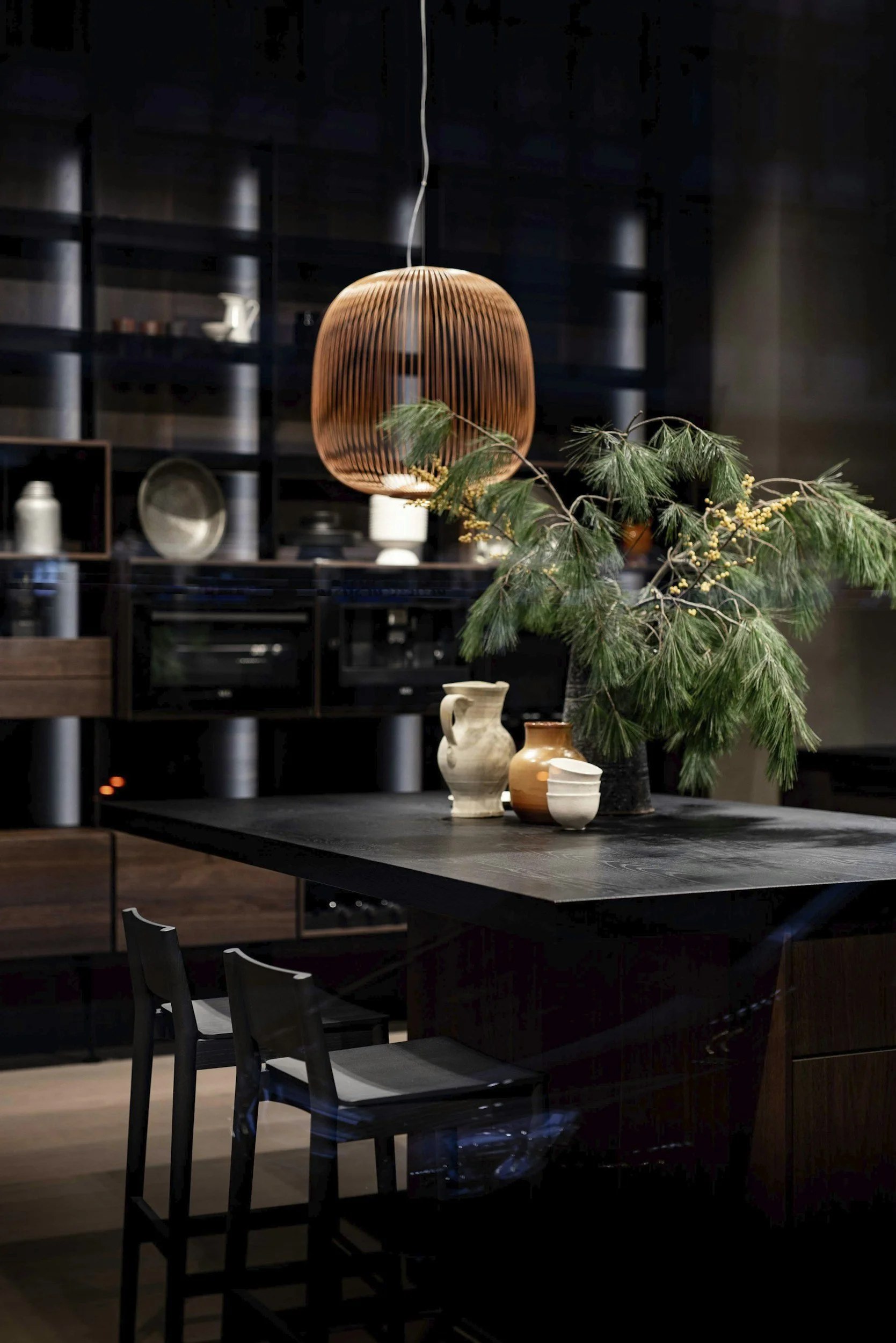 A modern dining room with a dark wooden table, black chairs, decorative vases, a large green tree, and a wooden pendant light hanging from the ceiling.