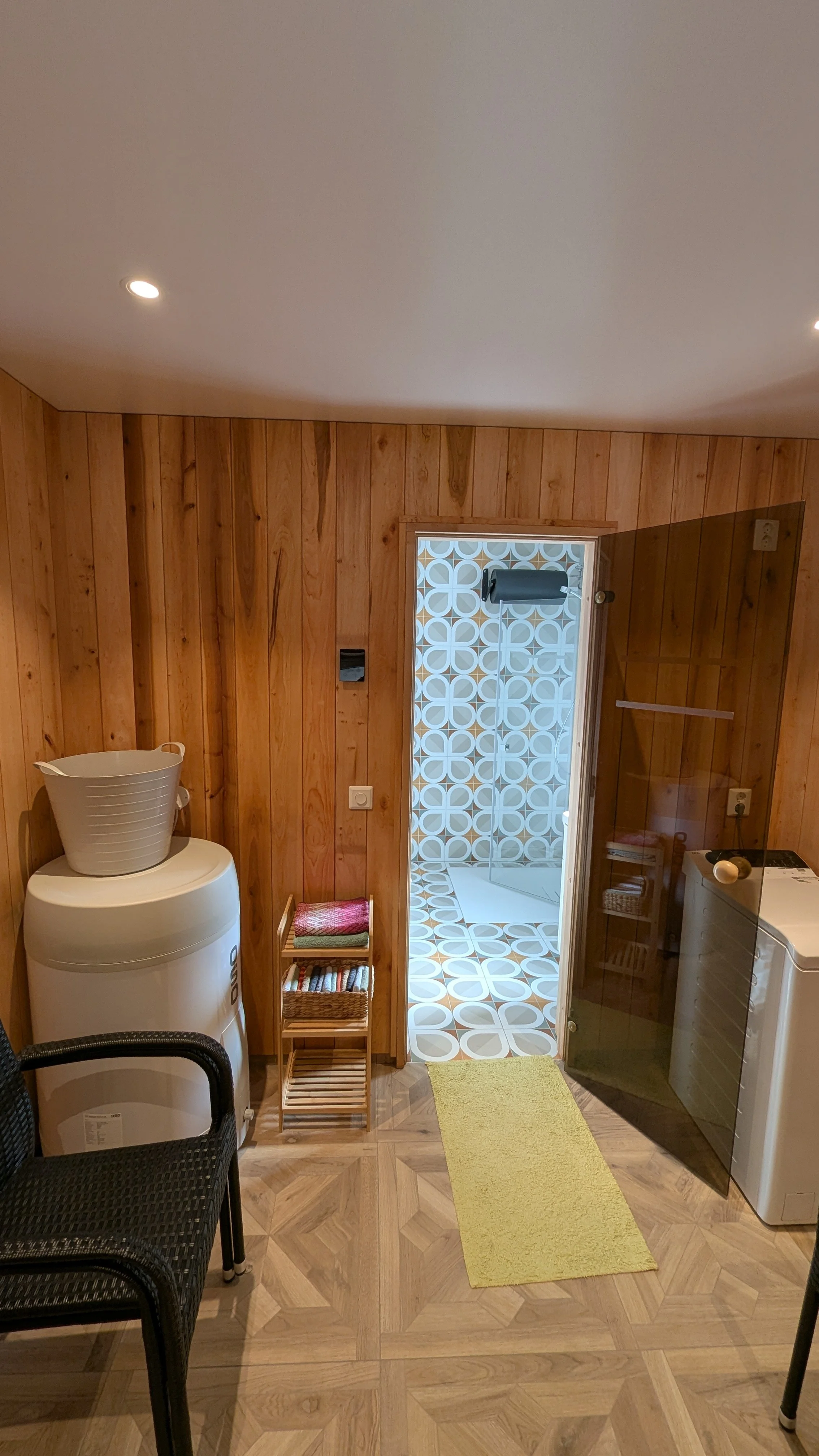 Et rom med tre lamper i taket, tre grønne matter, en vaskemaskin, en liten hylle med vaskeboller, en gul teppe, og en inngang til et baderom med fliser. Veggene er dekket av trepanel.