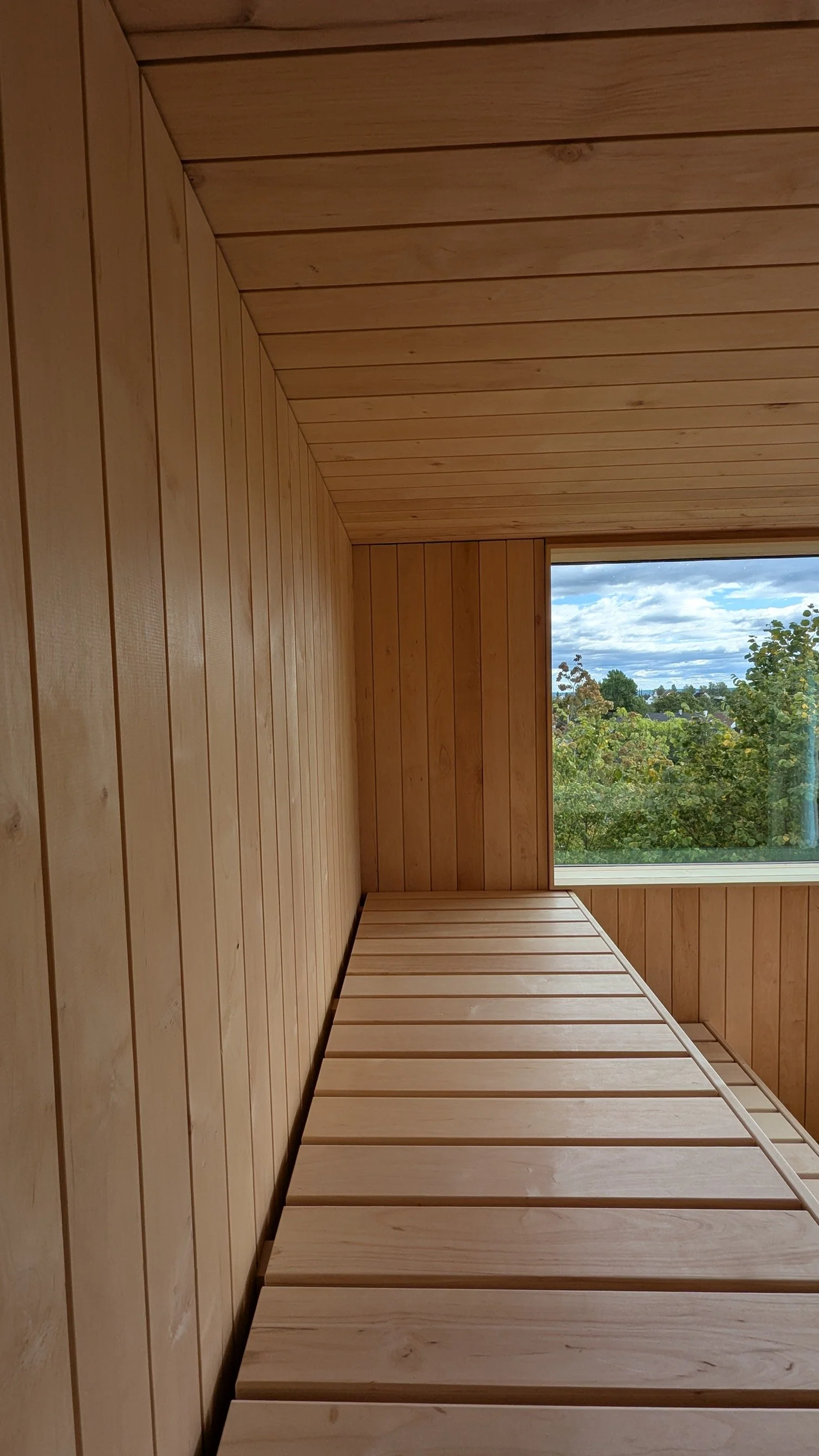 Innendørs bilde av en rom med trepanelvegger og tak, og et stort vindu som viser utsikt over trær og en overskyet himmel.
