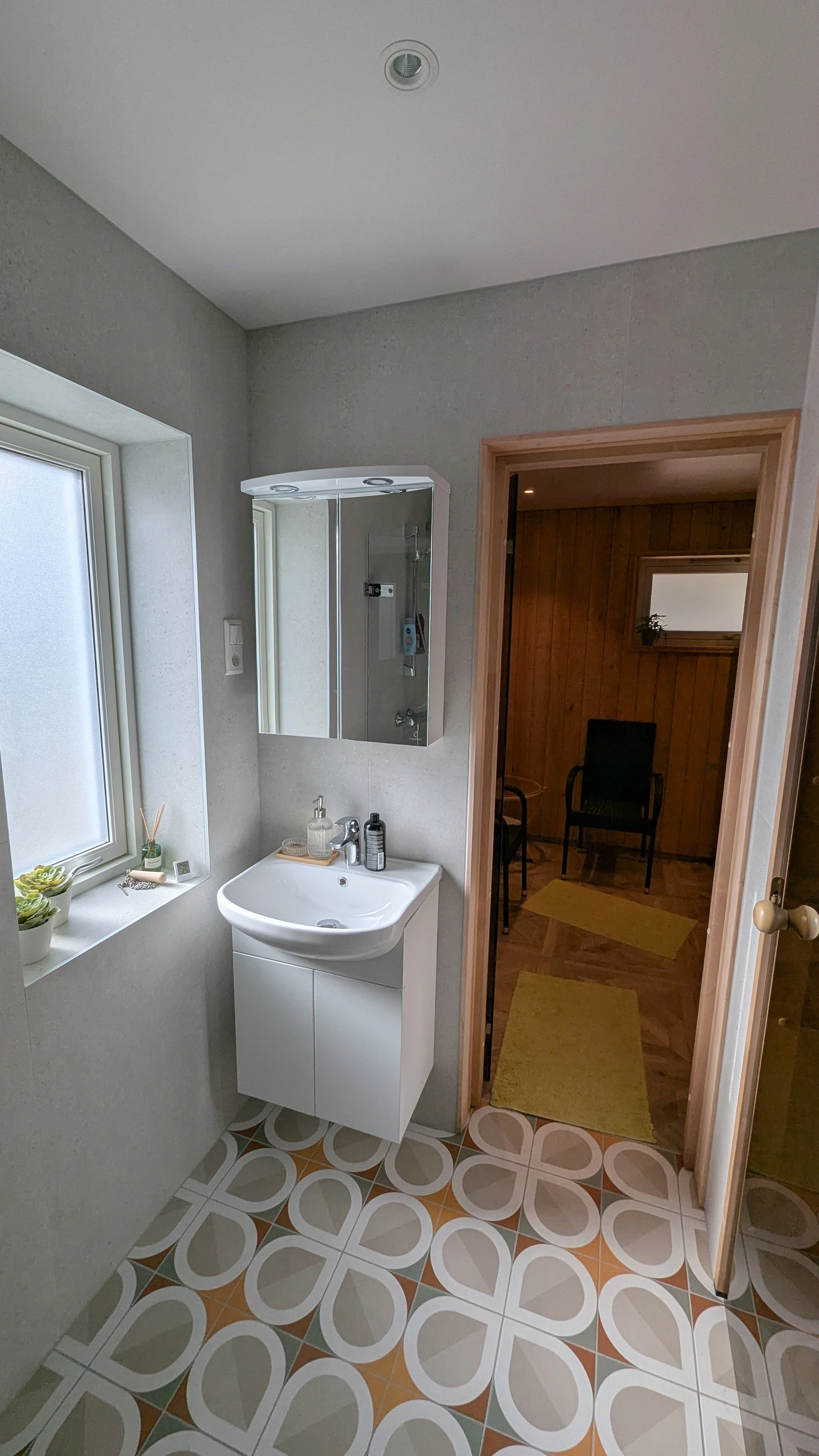 Badeværelse med hvid vask, spejl, vindue og et tilstødende rum med stol og gulvfliser med mønster.