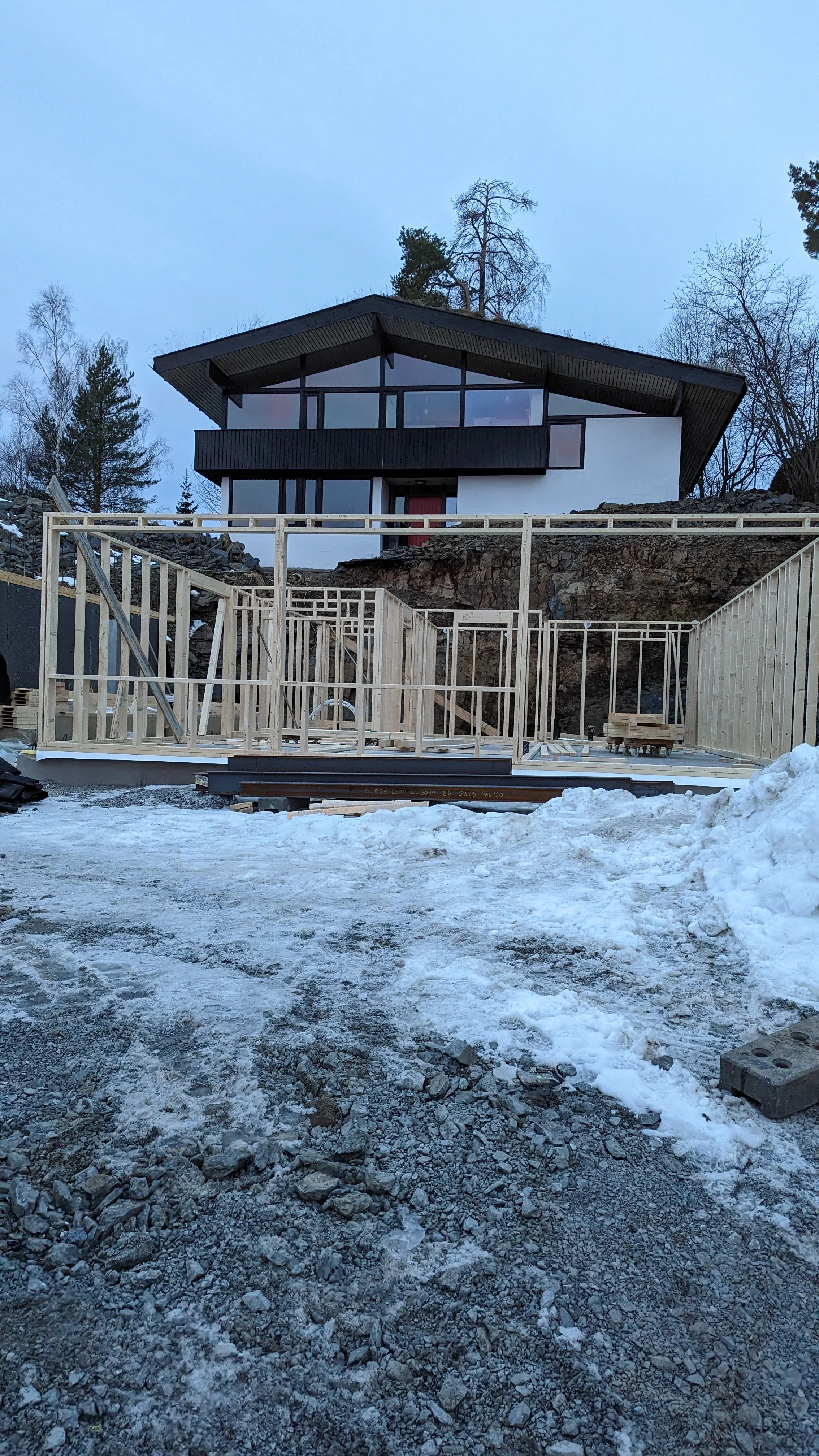 Et hus under bygging, med en ramme av tre og en hvit grunnmur, plassert på en snødekt tomt med trær i bakgrunnen.