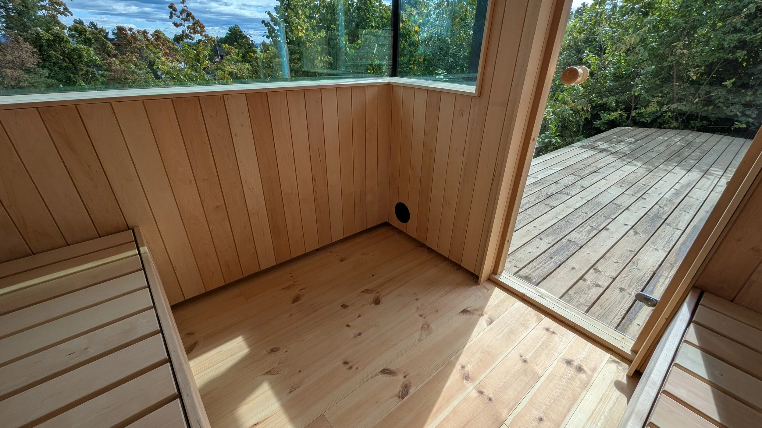 Innendørs utsikt over et lite rom med trepanel på vegger og gulv, en stor vindu mot et grønt utendørsområde, og en dør som leder ut til en treterrasse.