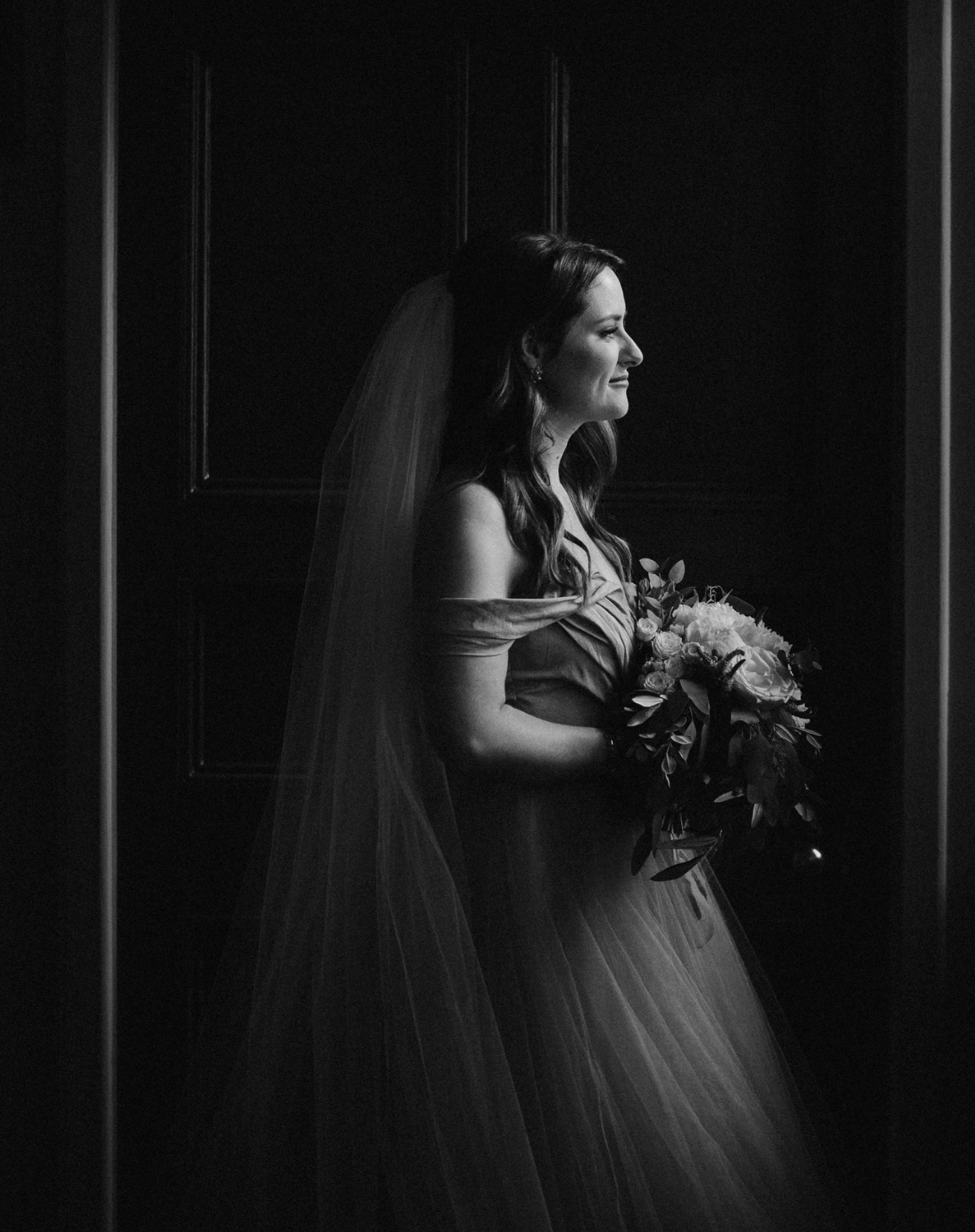 Black and white photo of a bride in a wedding dress holding a bouquet, standing sideways in front of a dark background, with her face in profile and smiling.