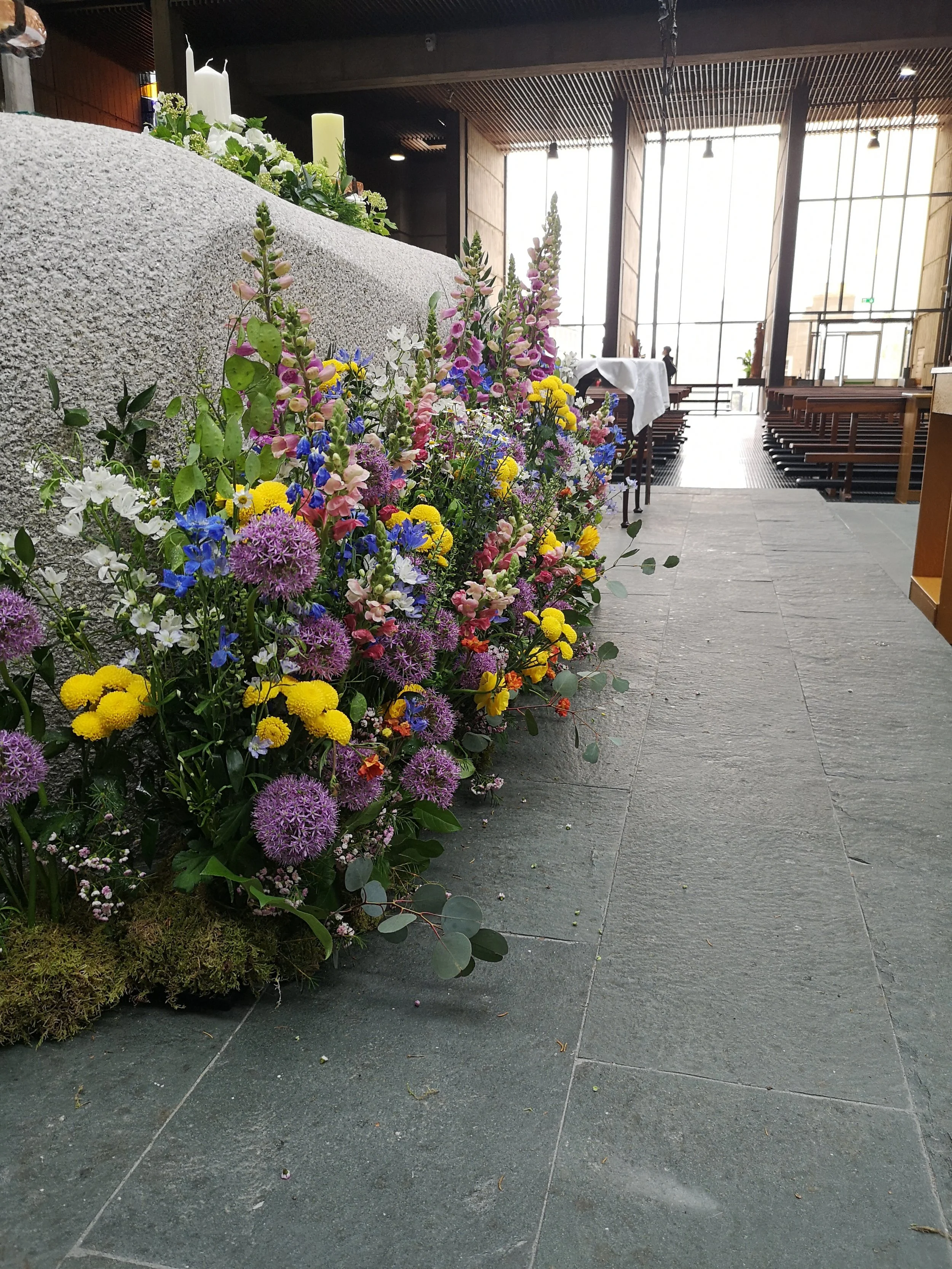 Colorful flower arrangement with various flowers, including yellow, purple, pink, and white, next to a textured stone wall inside a modern building with large windows and wooden ceiling.
