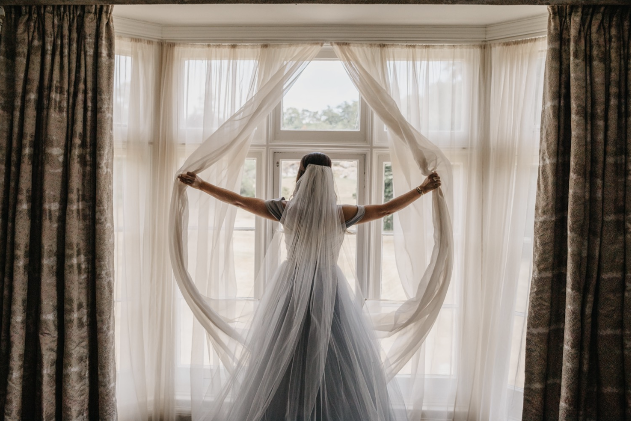 Bride with long veil and white gown standing in front of large window, holding open curtains with both arms.