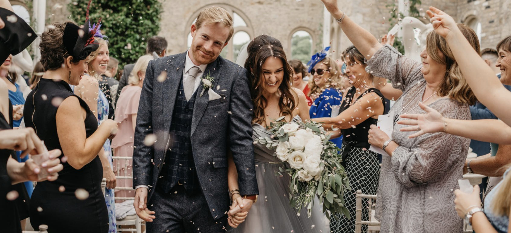 A newly married couple walking down the aisle at their wedding, holding hands, while surrounded by smiling guests celebrating with confetti.