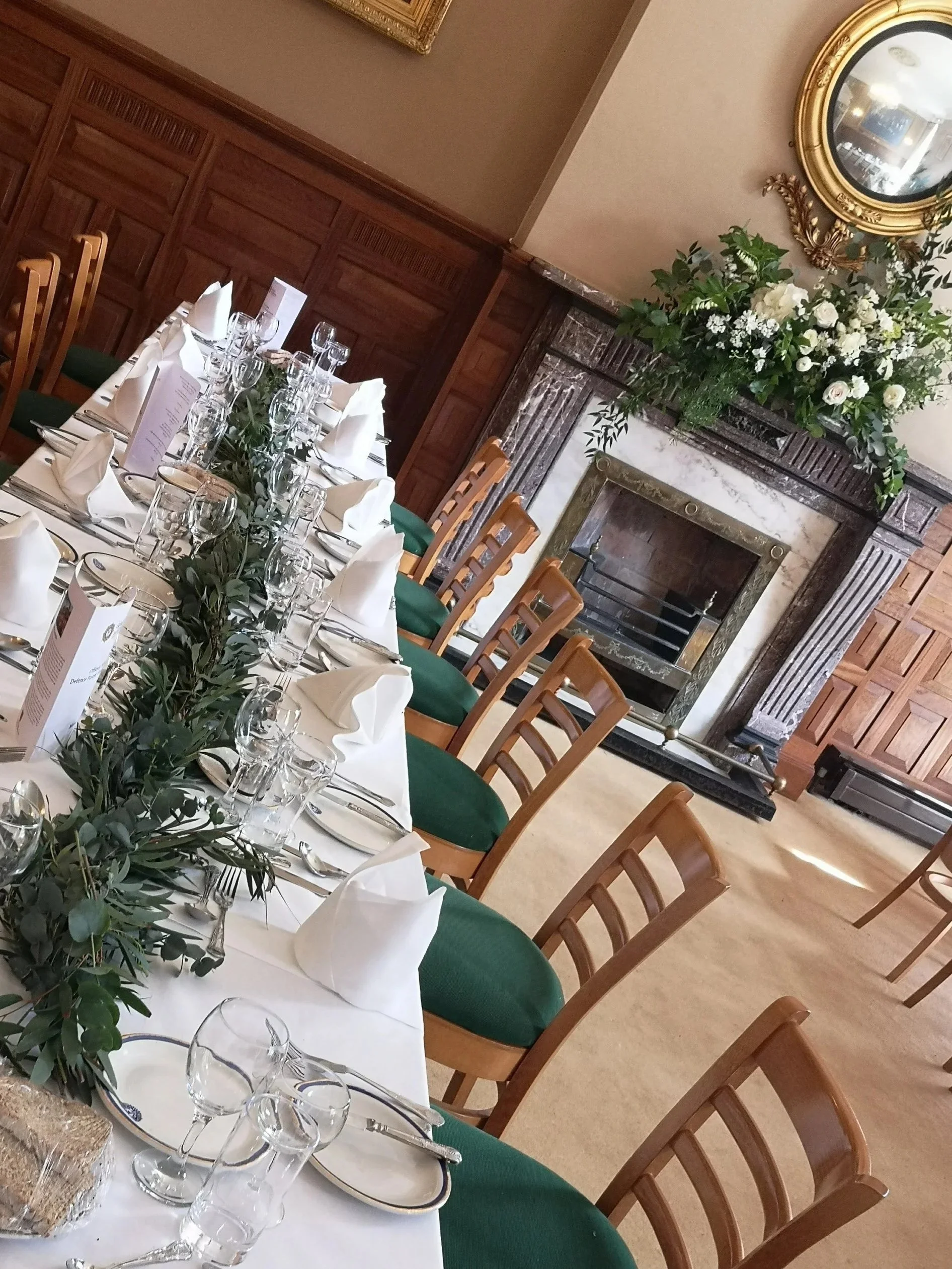 Elegant dining table set for a formal event with white tablecloths, white napkins, glassware, and green centerpiece foliage, in a room with wood-paneled walls, a fireplace with floral arrangement, and a gold-framed mirror.