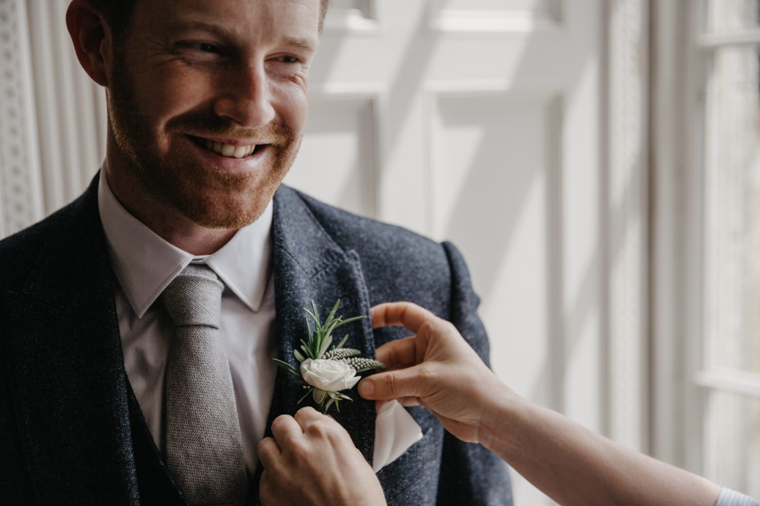 A man in a suit smiling as someone pins a white flower boutonniere onto his jacket lapel.