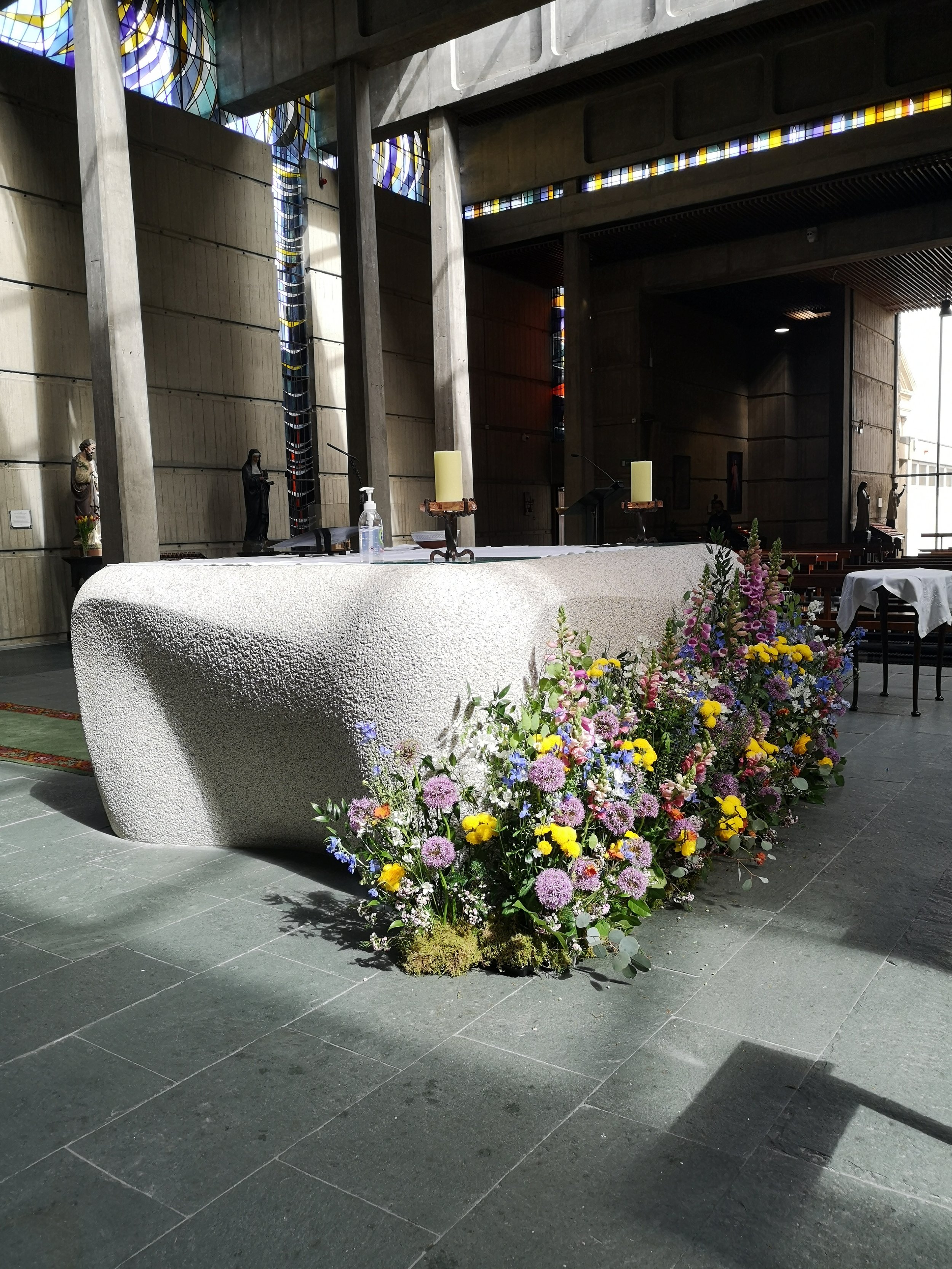 Interior of a church with a stone altar decorated with colorful flowers, candles, and a soap bottle, with statues and stained glass windows in the background.