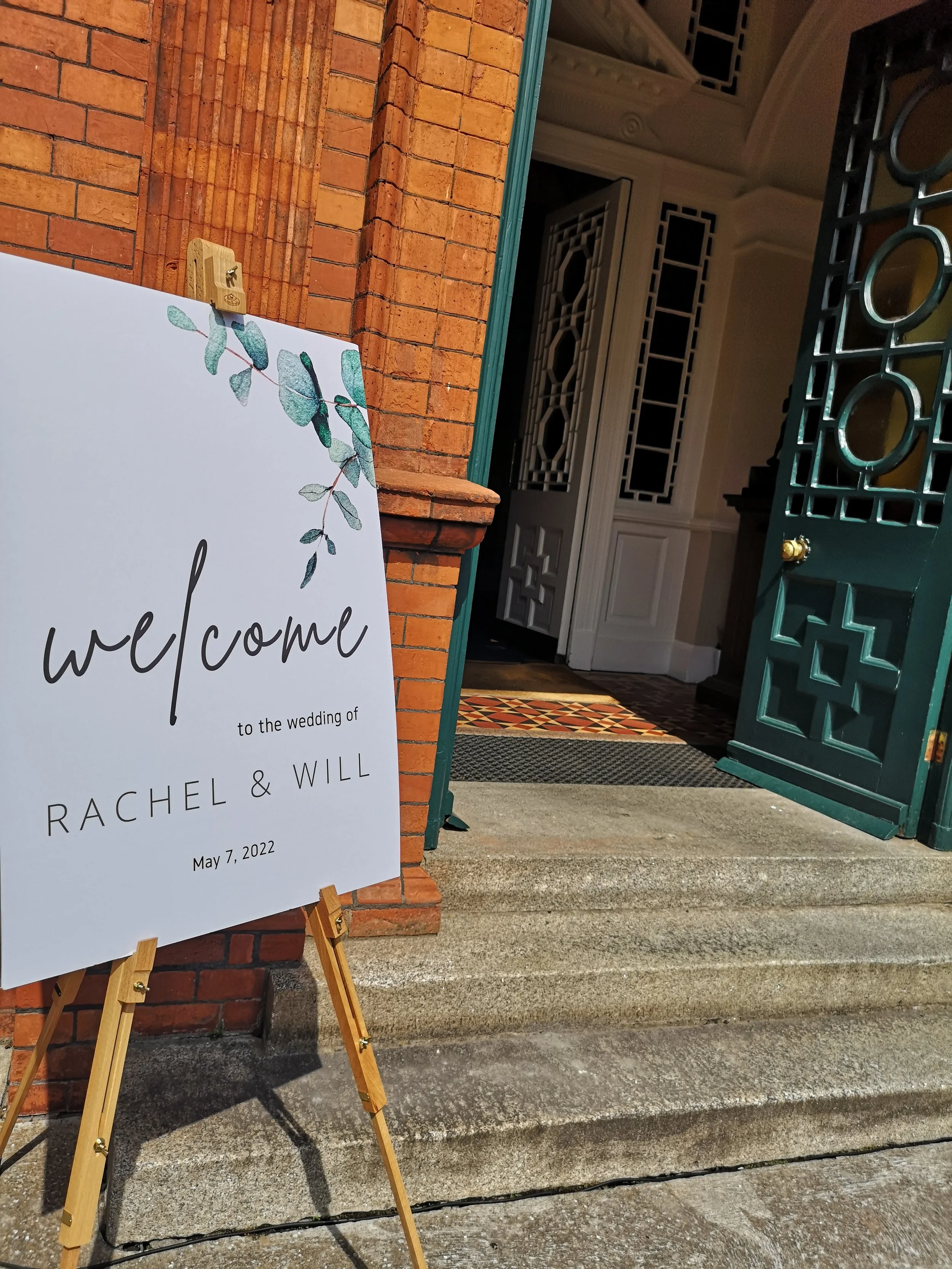 Wedding welcome sign placed on a wooden easel at the entrance of a building with an open door, brick and white walls, and a patterned rug inside. The sign has a eucalyptus branch illustration and reads: 'welcome to the wedding of Rachel & Will, May 7