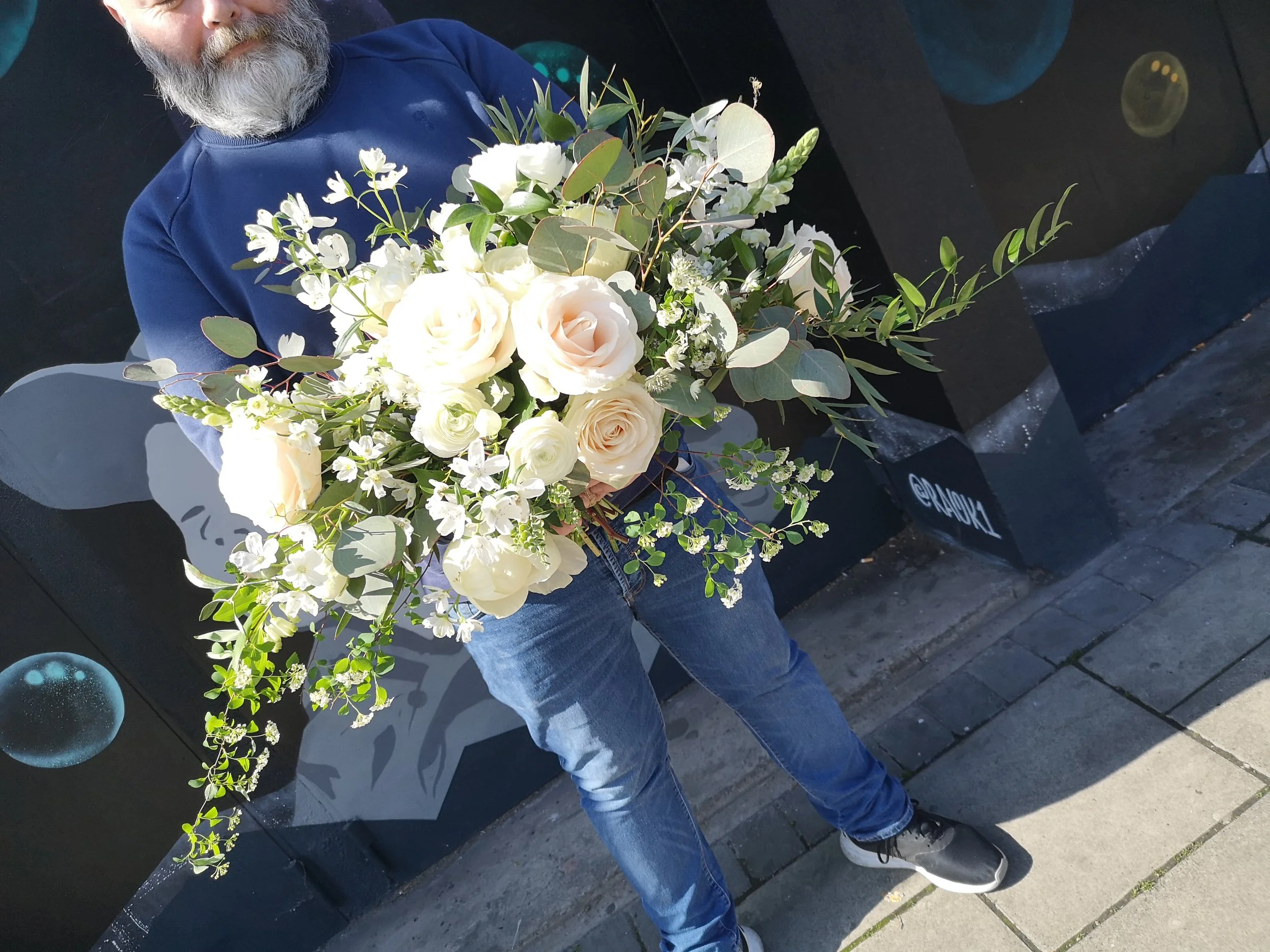 Person holding a large bouquet of white roses and greenery in front of a black wall with artistic circles and text.