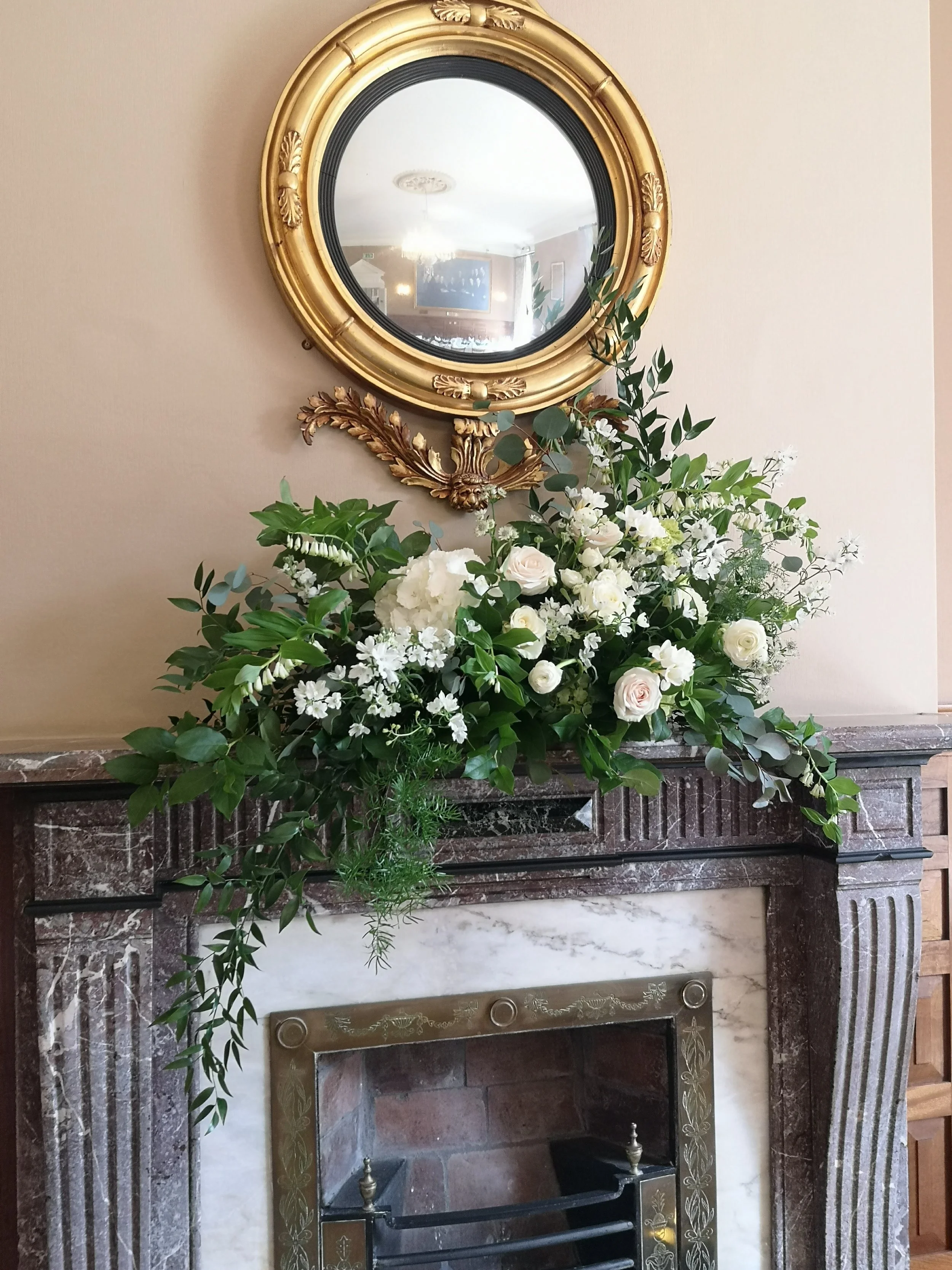 A decorative fireplace mantel with a bouquet of white and green flowers and leaves, topped with a round gold-framed mirror.