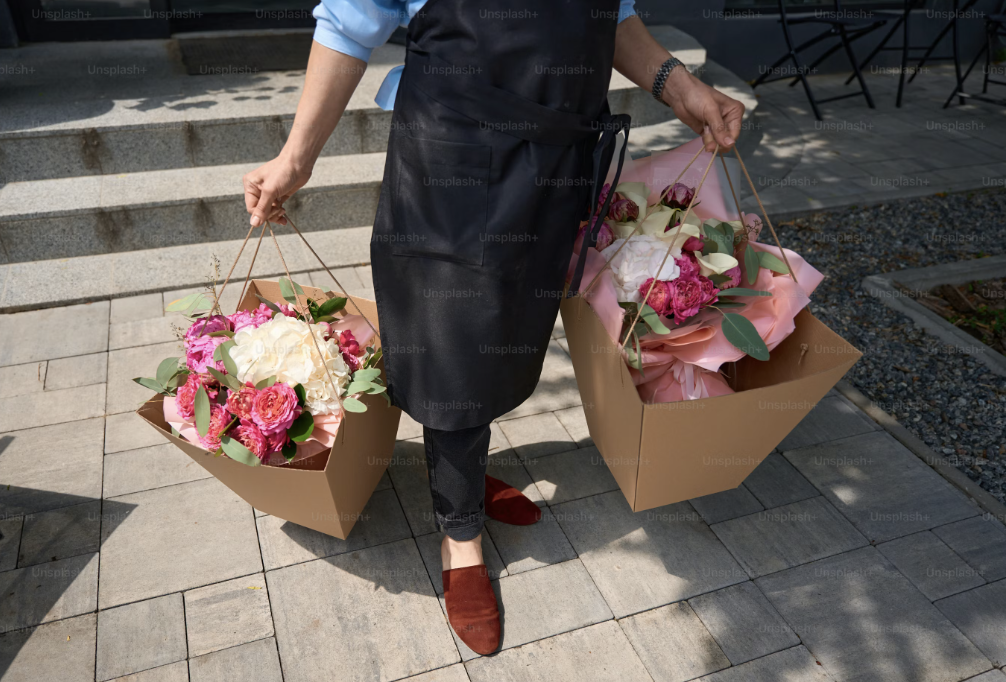 Person standing outdoors holding two large boxes of pink and white flowers with green leaves, wearing a black apron, blue shirt, black pants, and red shoes.