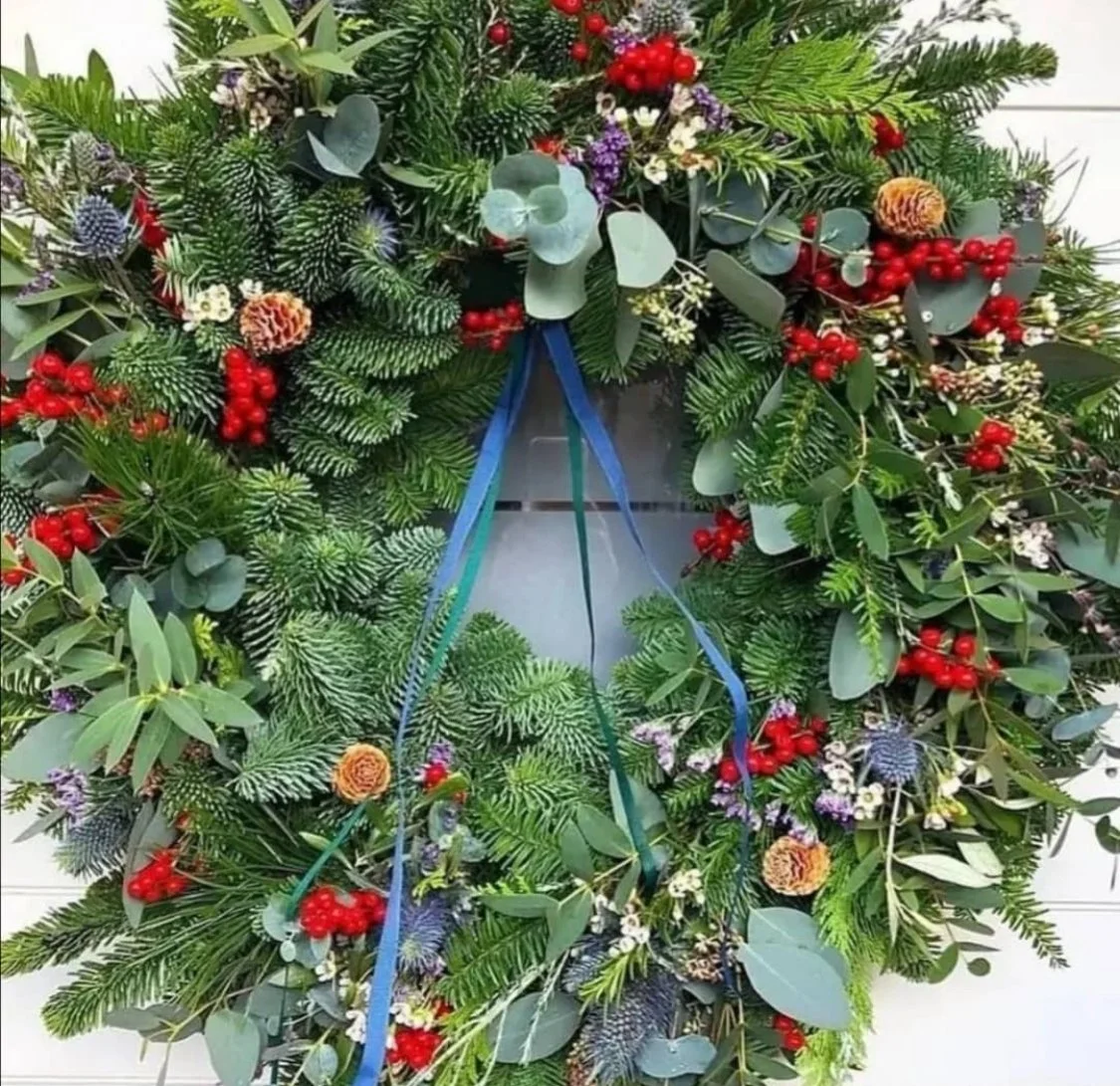 A Christmas wreath decorated with various green foliage, berries, pinecones, and small flowers, with blue ribbon loops at the top for hanging.
