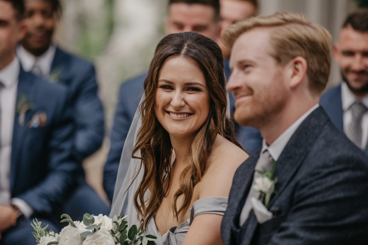 A bride and groom smiling at each other during a wedding ceremony, with wedding party members in the background.