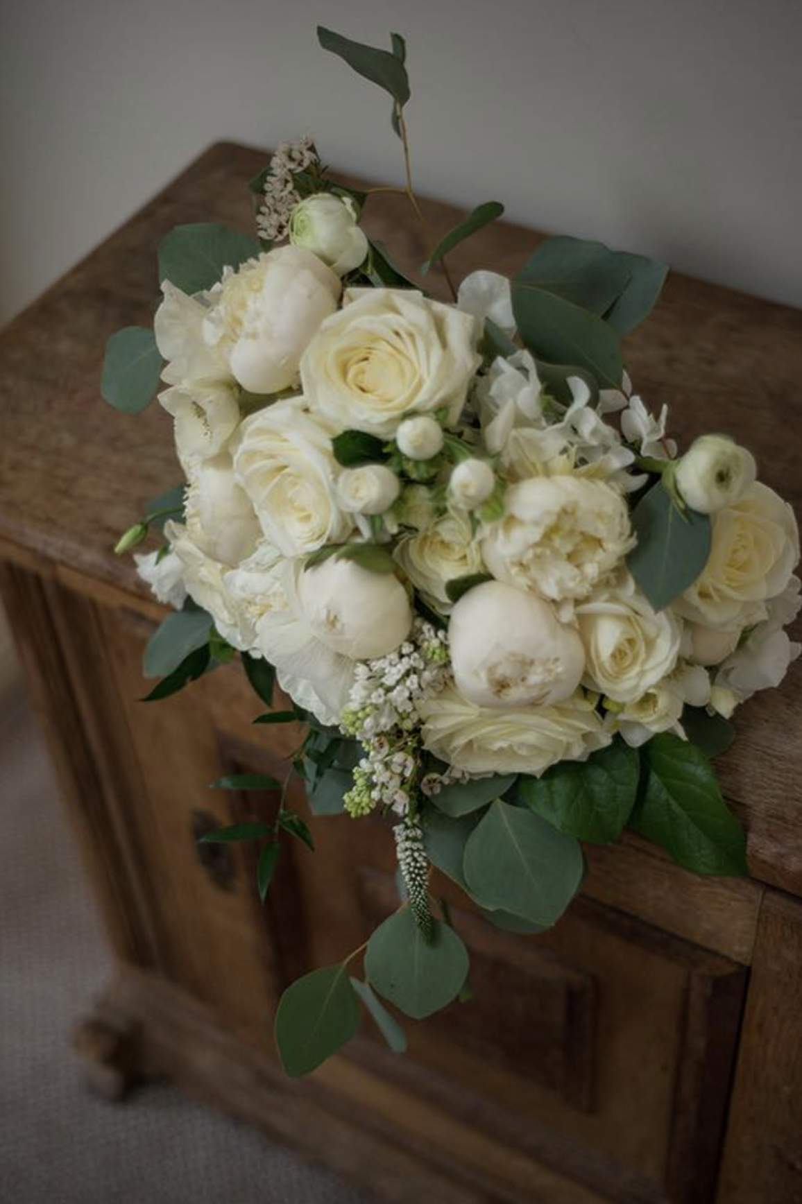 A bouquet of white roses, peonies, and other white flowers with green leaves resting on a wooden surface.