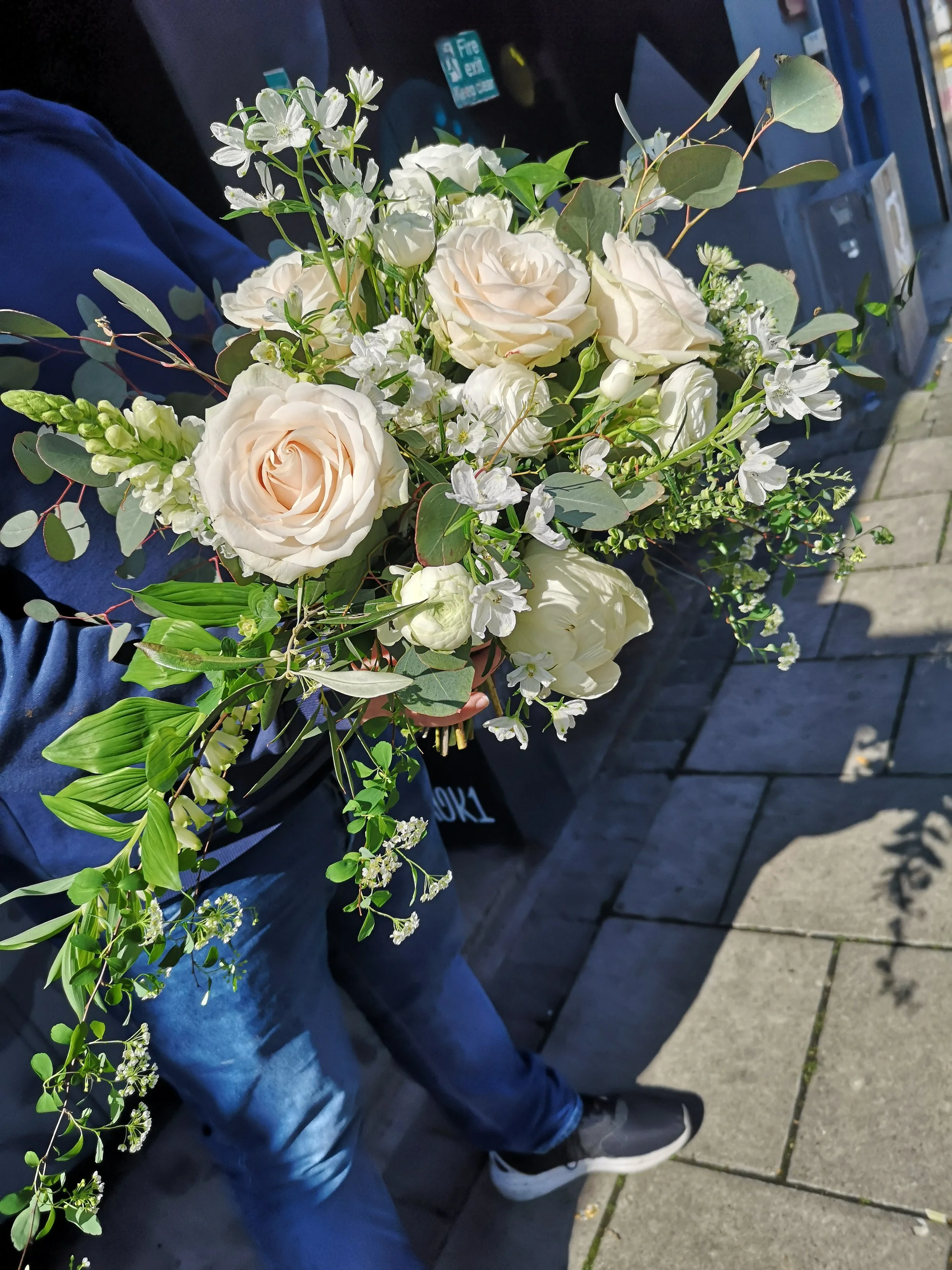 A person holding a large bouquet of white and cream-colored roses, white smaller flowers, and green foliage, standing outside on a sidewalk.