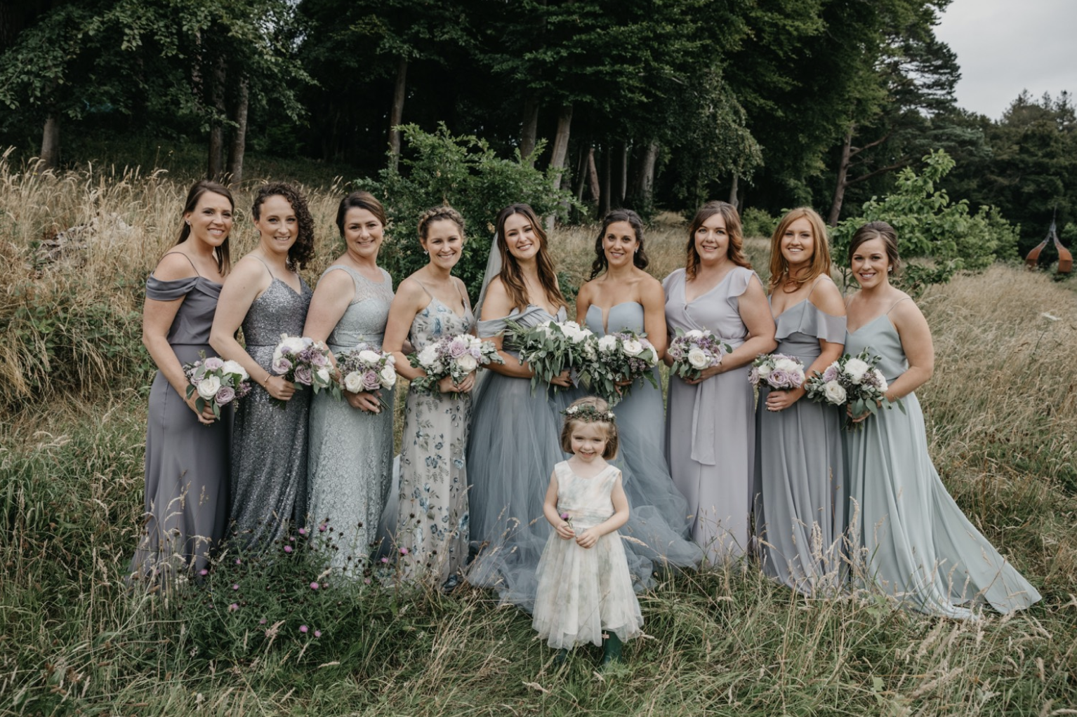 A bride and her flower girls and bridesmaids posing outdoors in a natural setting with trees and tall grass, all holding bouquets of flowers. The bride is in the center wearing a wedding gown, and a young girl in a floral dress is in front of her.