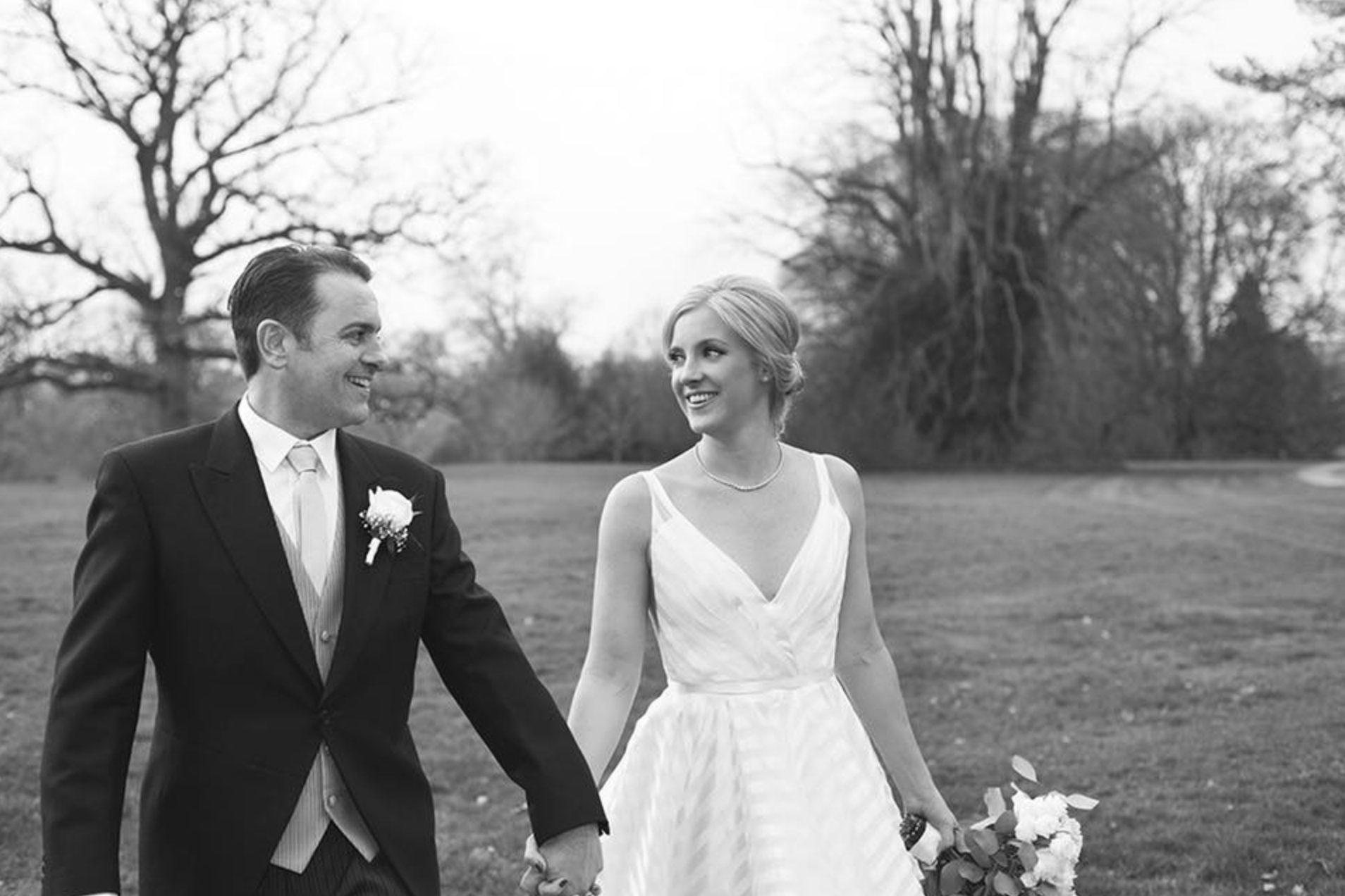 A black and white photo of a bride and groom holding hands outside, smiling at each other. The groom is dressed in a suit with a boutonniere, and the bride is in a white wedding dress holding a bouquet of flowers, with trees in the background.