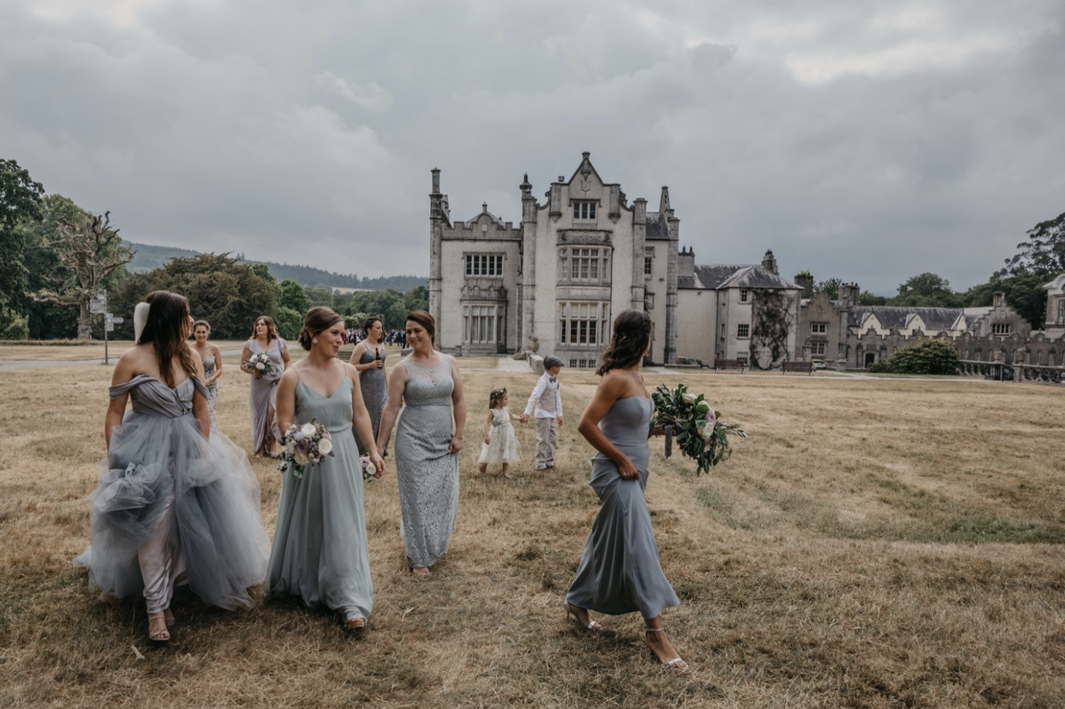 Bridal party walking on grass outside of a castle-like building, some women holding bouquets, children walking nearby, overcast sky.