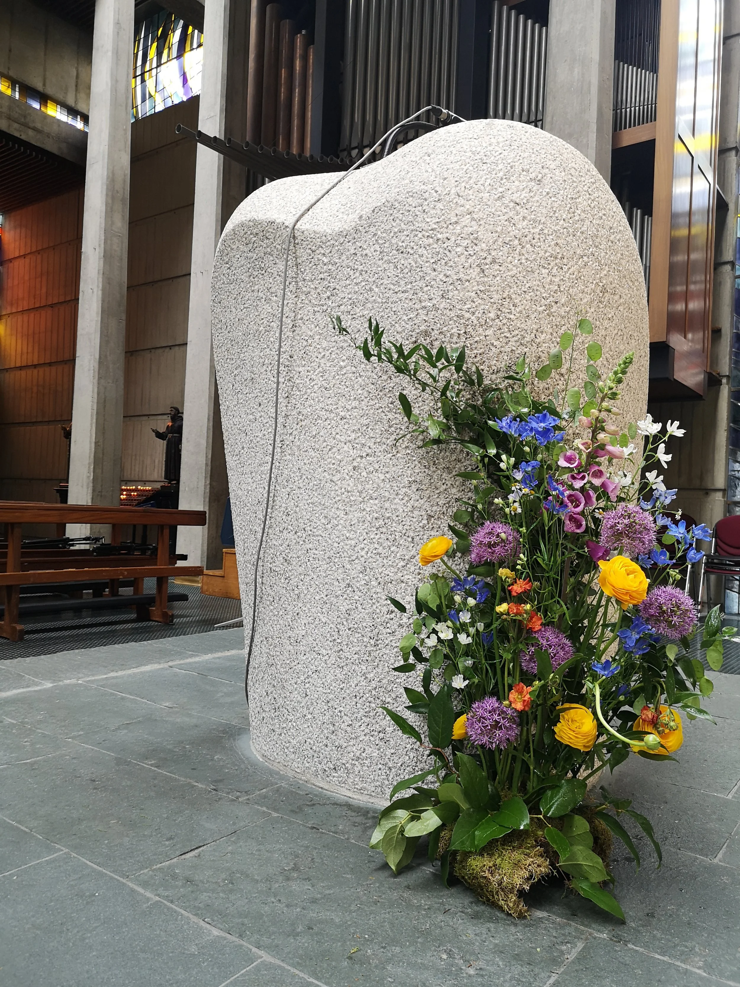 A large stone monument with a rounded top, adorned with a colorful floral arrangement at its base, inside a building with concrete columns and wooden accents.