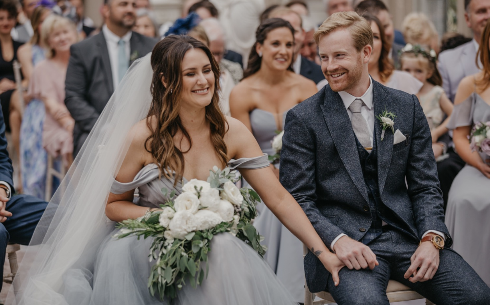 A bride and groom sitting together during their wedding ceremony, holding hands and smiling. The bride is wearing a strapless wedding dress with a veil, holding a bouquet of white flowers and greenery. The groom is dressed in a dark gray suit with a 