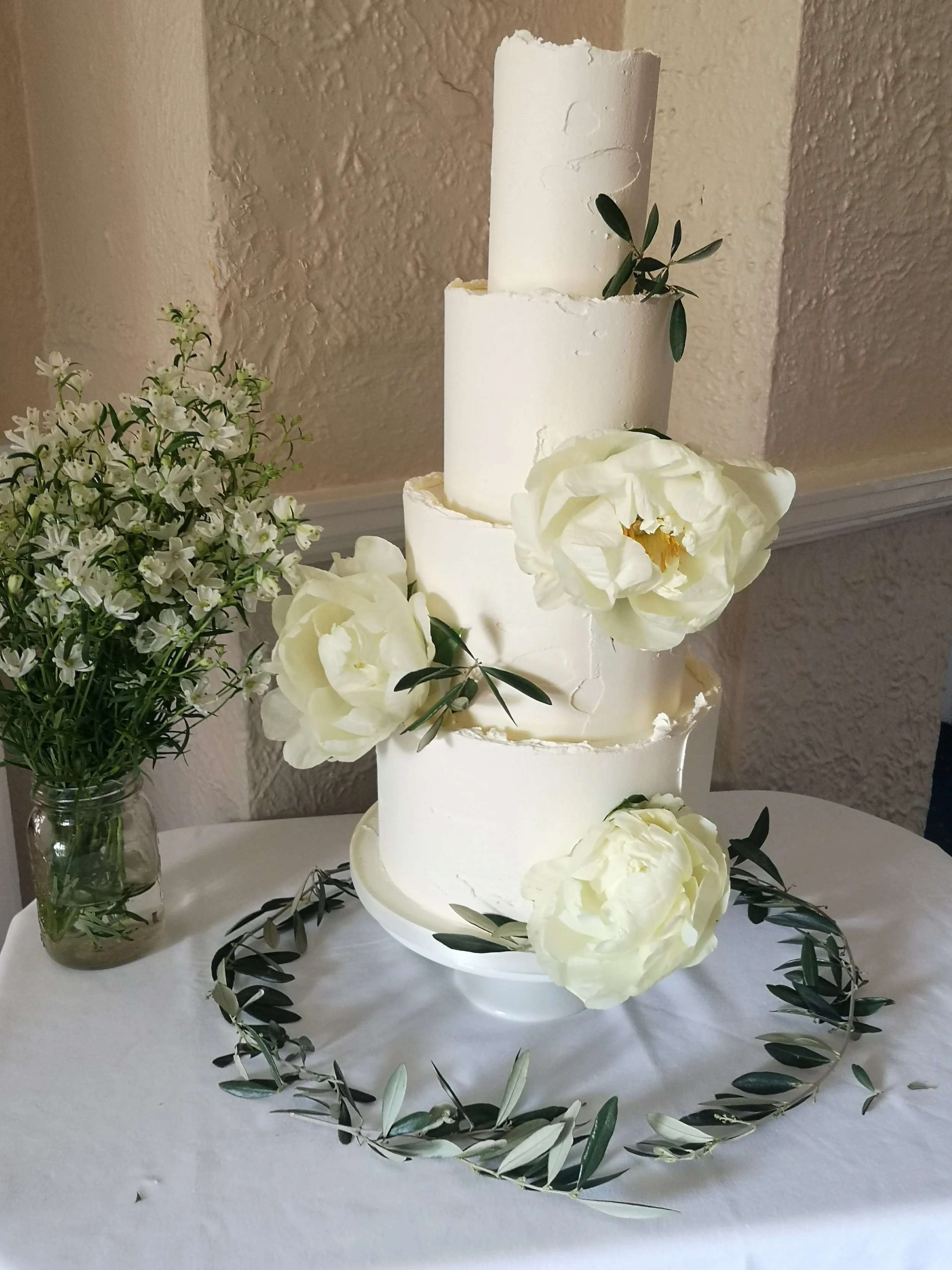 A four-tier white wedding cake decorated with large white flowers and green leaves, placed on a white table with a garland of greenery around the base, next to a vase of white flowers, against a beige textured wall.