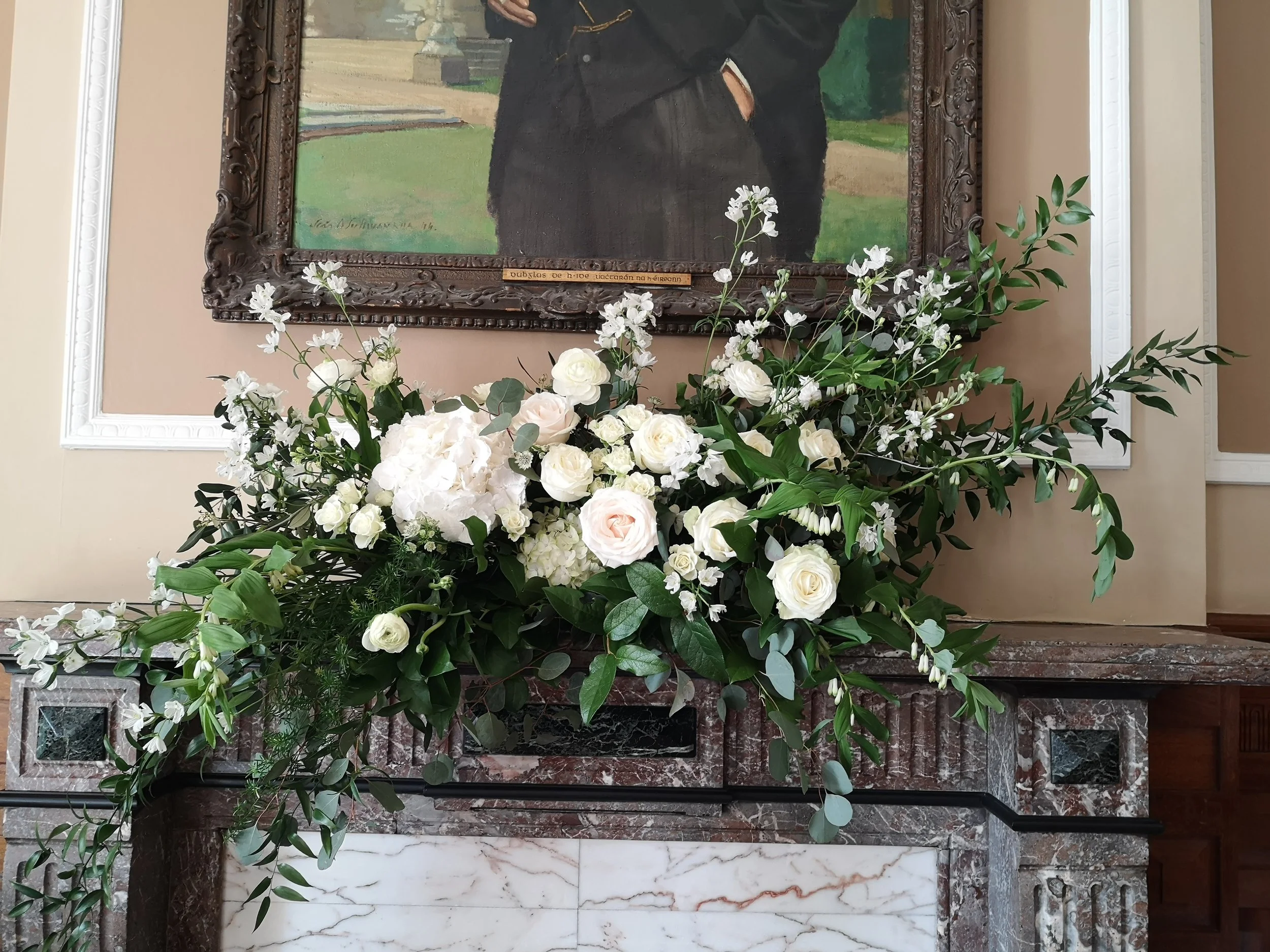 A large bouquet of white flowers and green foliage on a marble and stone mantelpiece, with a framed portrait of a person in a dark suit hanging above.