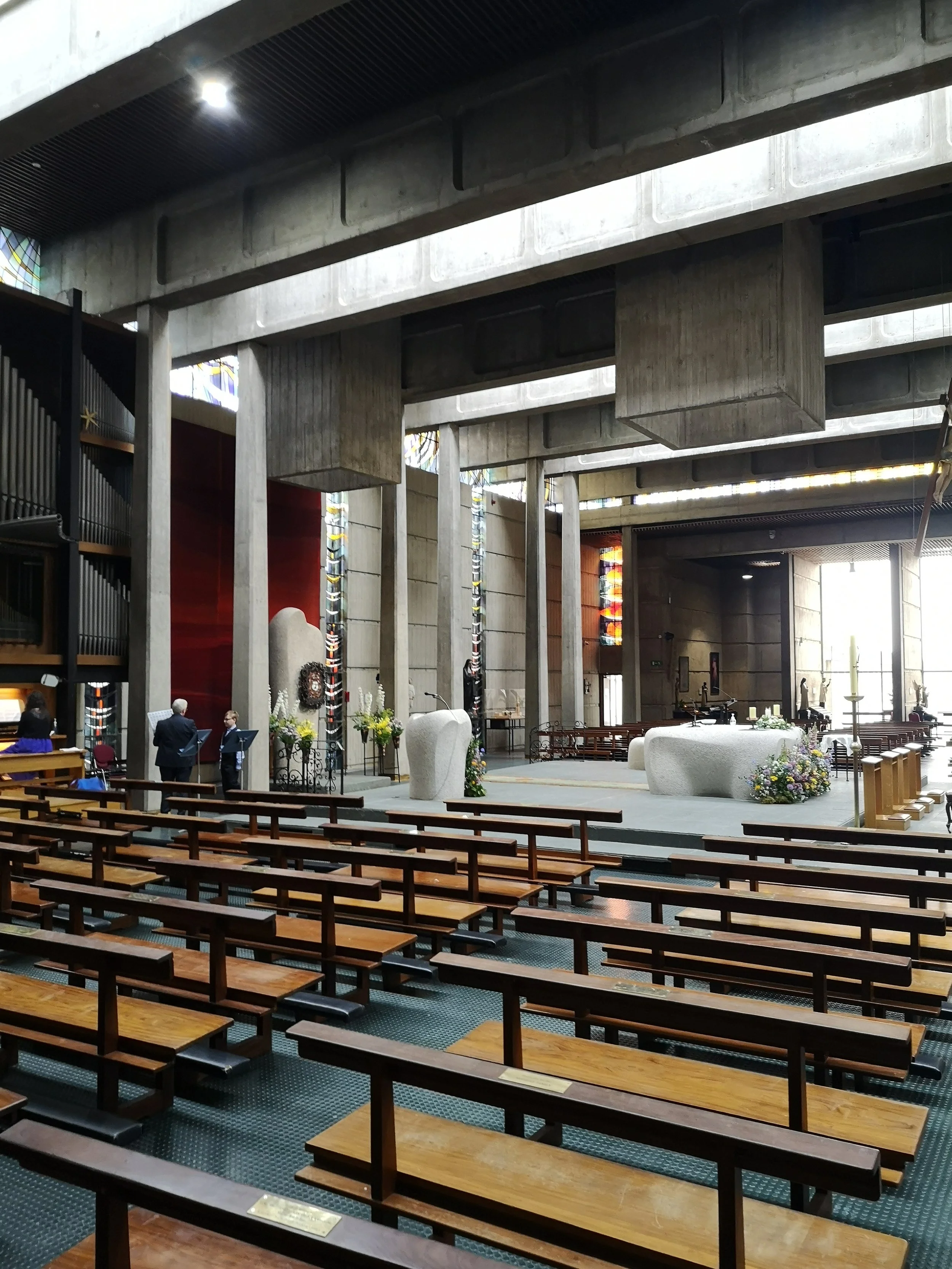 Interior of a church with wooden pews, a raised altar with floral arrangements, and stained glass windows, during a daytime service or ceremony.
