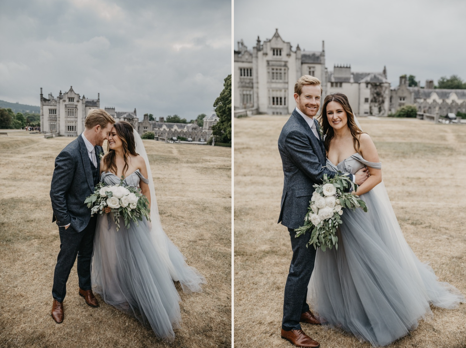 A smiling bride and groom standing close together on a grassy field in front of a large historic castle, with the groom wearing a blue tweed suit and the bride in a flowing gray tulle gown holding a large bouquet of white flowers and greenery.
